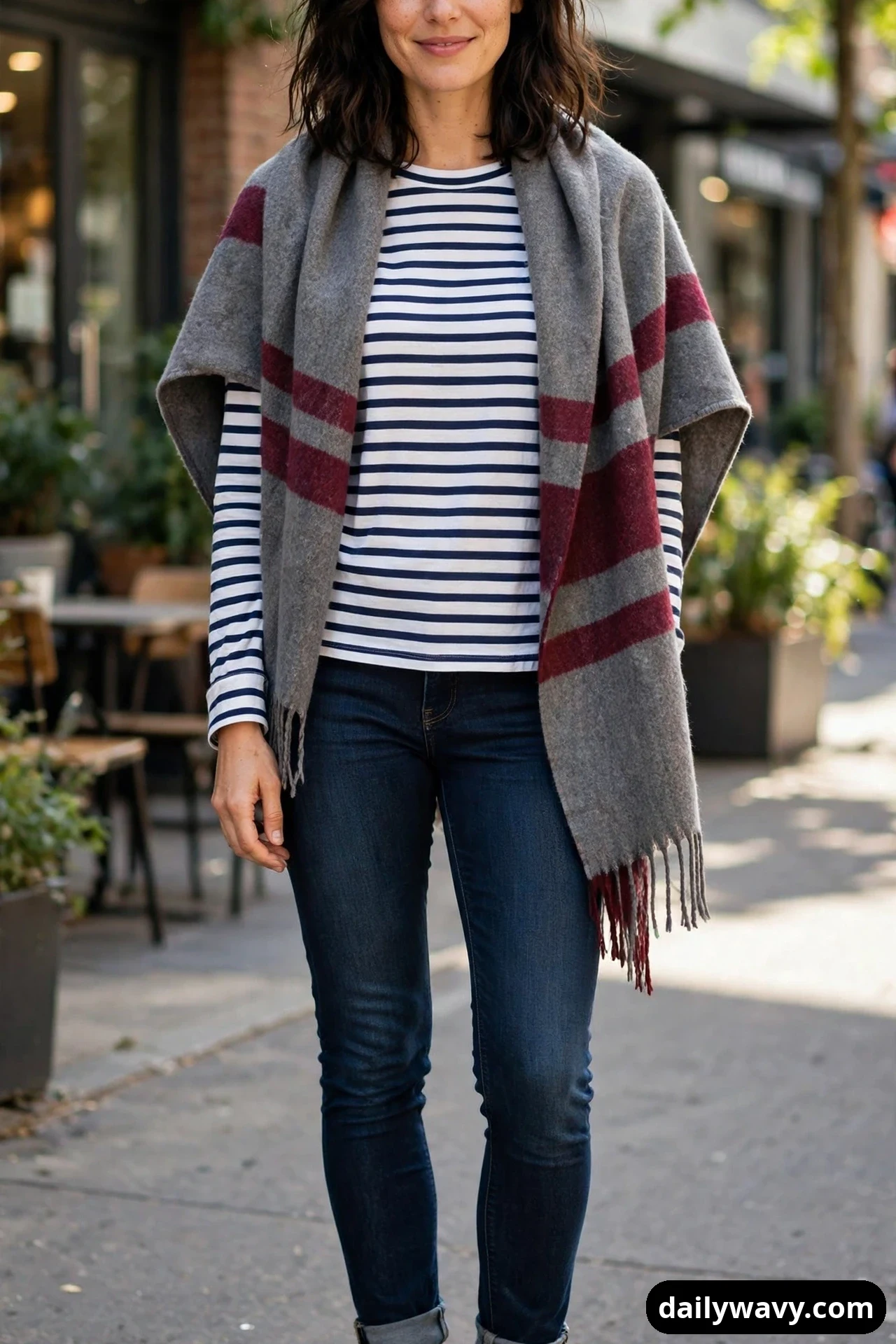 A woman wears a navy-and-white striped long-sleeve tee with dark skinny jeans, and a grey-and-burgundy striped blanket scarf draped open over her shoulders.