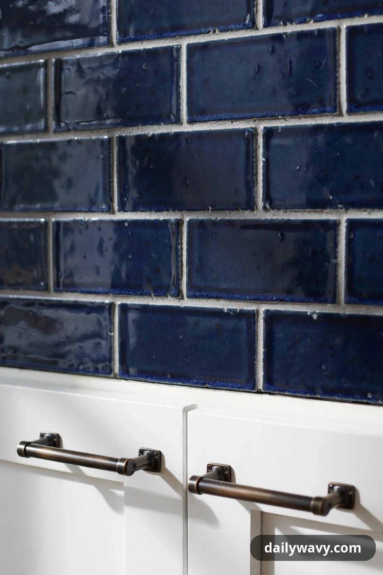 A close-up shows glossy deep navy subway tiles with light gray grout above white cabinet doors fitted with dark bronze bar pulls.