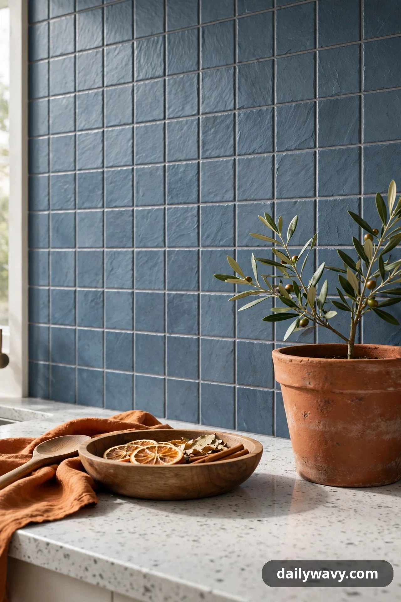 Textured slate-blue square tiles form the backsplash behind a counter styled with a terracotta pot holding an olive plant and a wooden bowl.