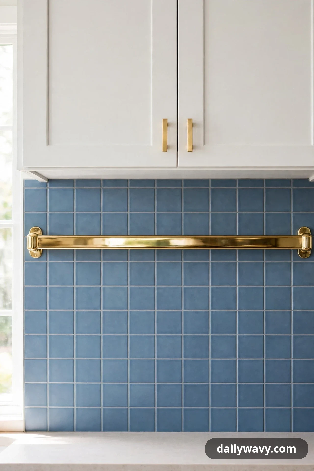 Matte steel-blue square tiles cover a backsplash wall crossed by a single polished brass rail below white cabinets.
