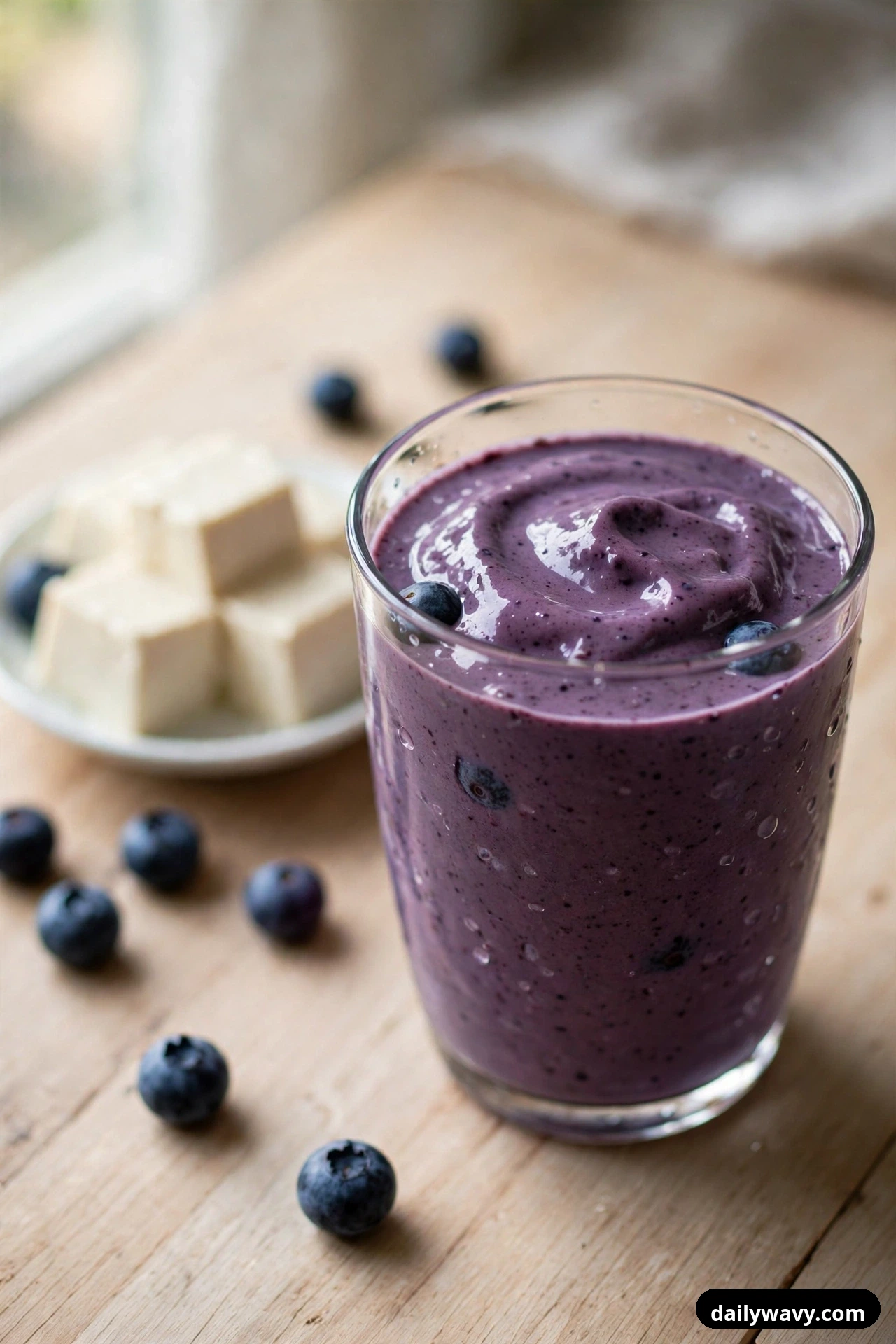 A closer view shows a glass of thick purple blueberry smoothie with a swirled top, with a small plate of tofu cubes and scattered blueberries behind it on a light wood table.