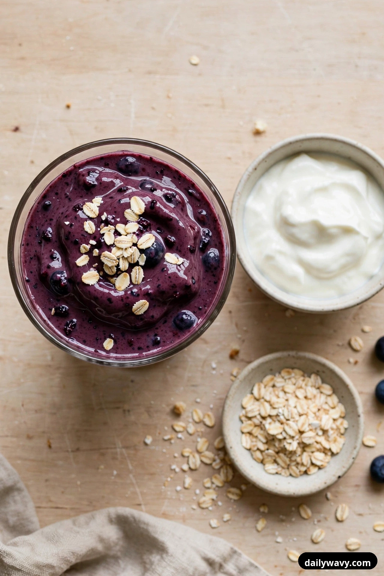 Seen from above, a glass of dark purple blueberry smoothie topped with rolled oats sits beside a bowl of Greek yogurt and a small dish of rolled oats on a pale wooden surface.