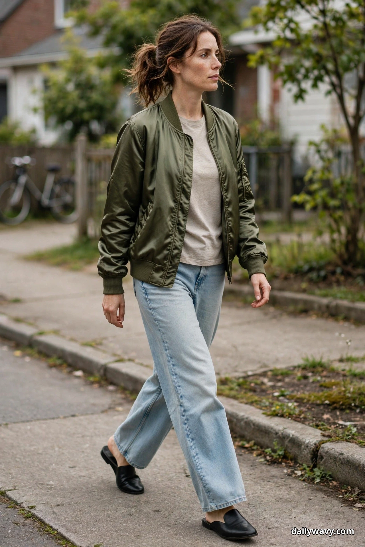 A woman walks along a residential sidewalk wearing an olive satin bomber jacket open over an oatmeal tee, with light-wash wide-leg jeans and black flat mules