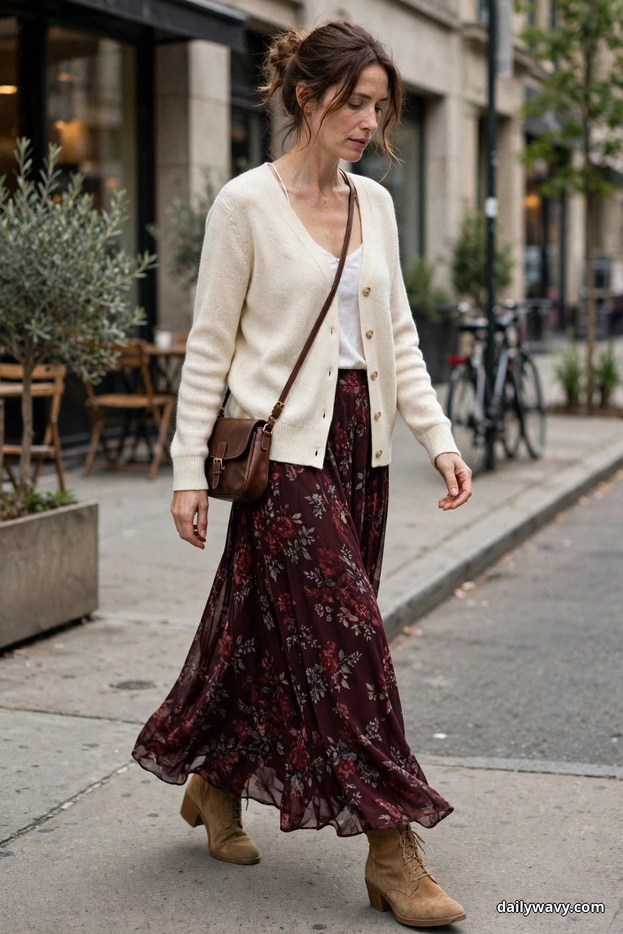 A woman walks along a city sidewalk wearing a cream buttoned cardigan open over a white camisole with a dark burgundy floral maxi skirt, a small brown leather crossbody bag and tan ankle boots
