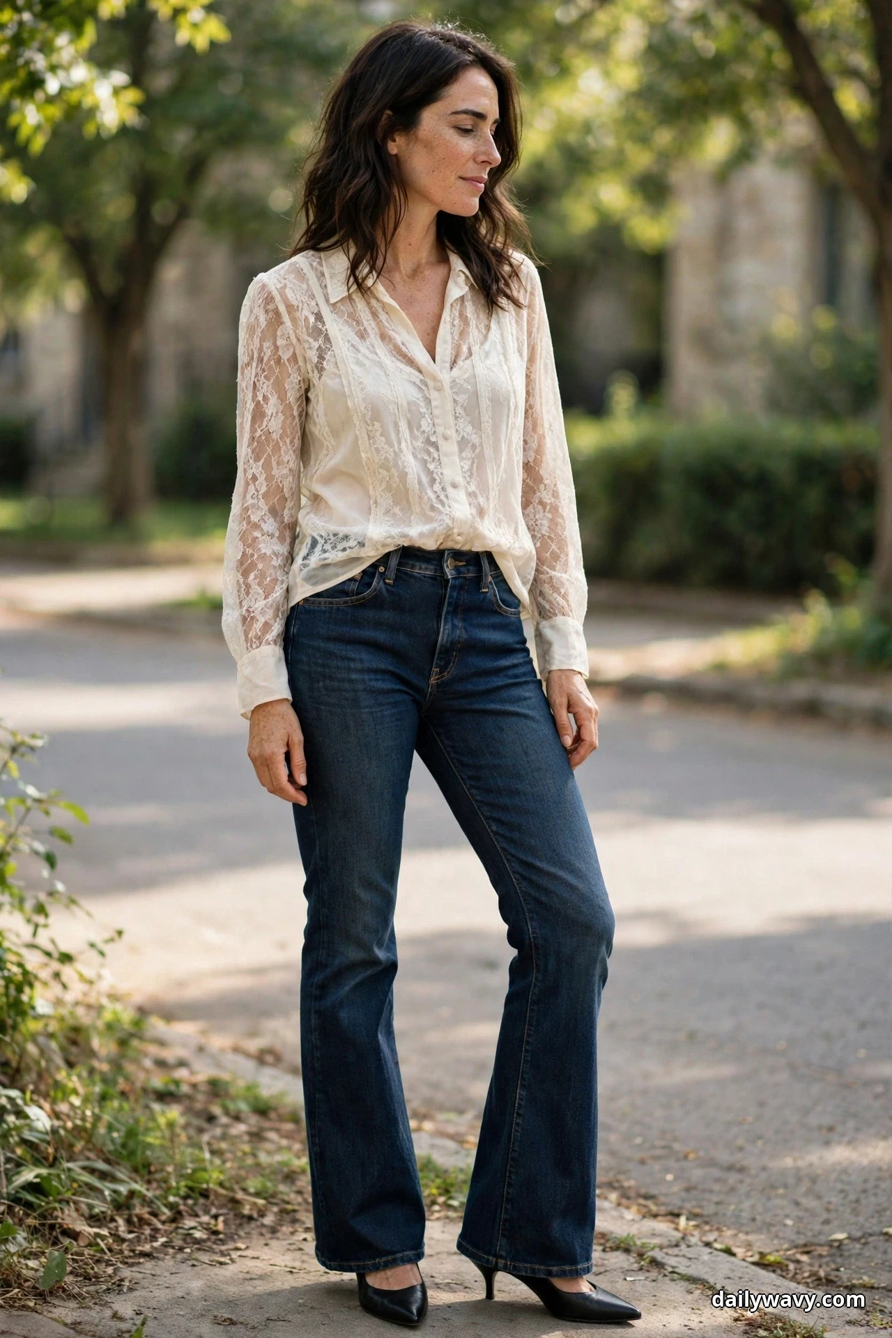 A woman stands outdoors wearing an ivory lace blouse with an opaque lining tucked into dark indigo bootcut jeans, with black pointed pumps showing under the flared hems