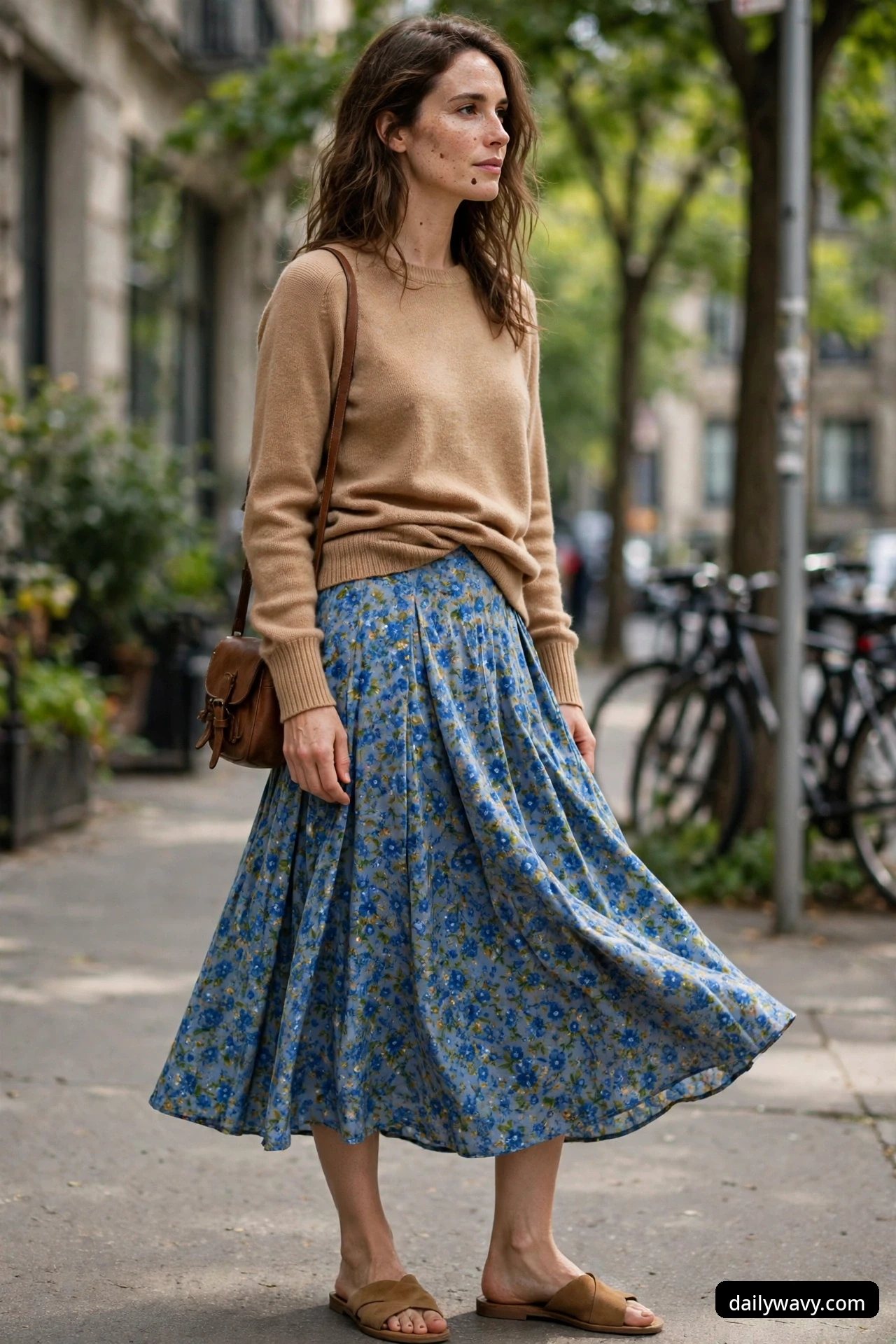 A woman stands on a sidewalk wearing a camel knit top tucked into a blue floral-print midi skirt, carrying a brown leather crossbody bag and tan slide sandals.