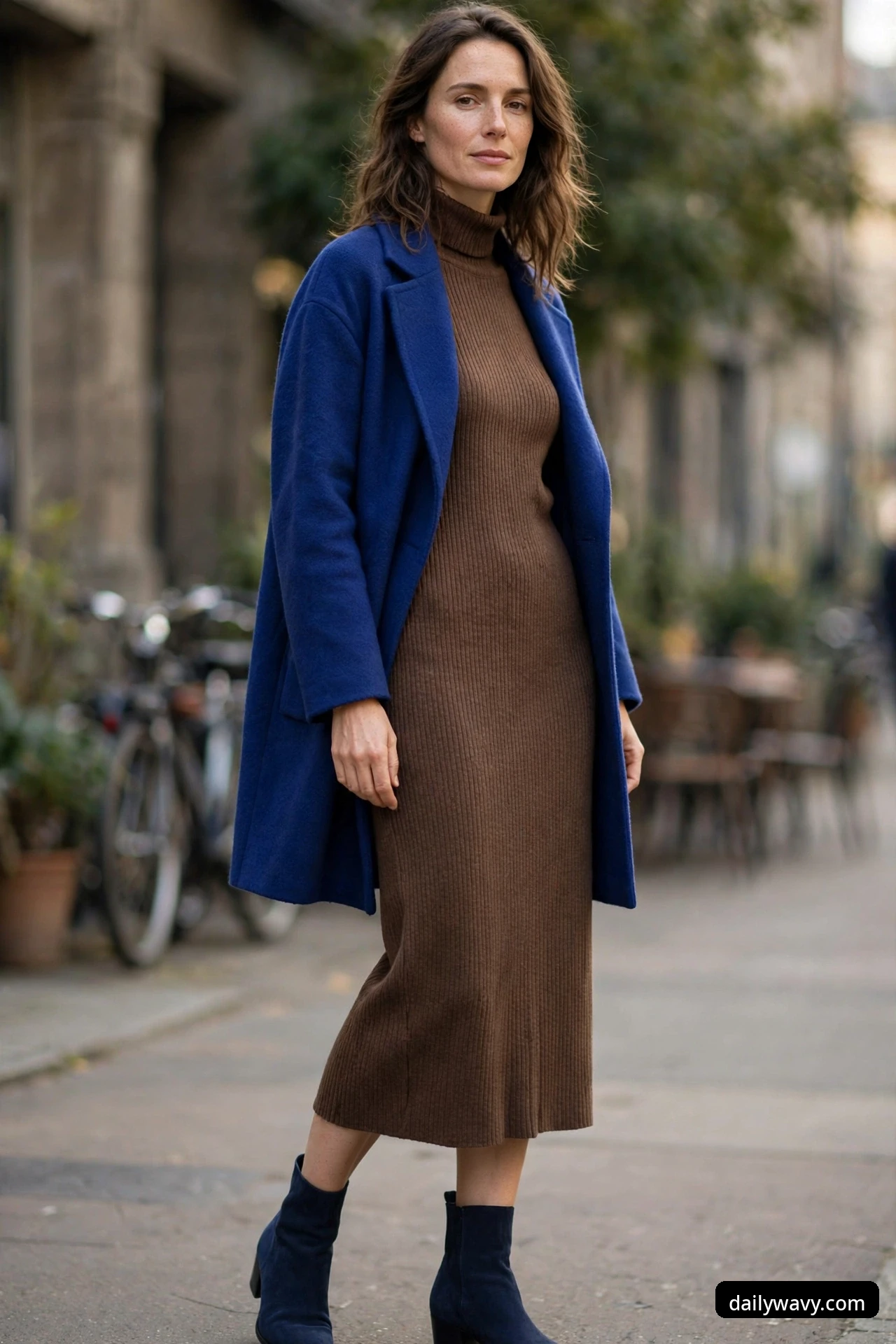 A woman walks along a sidewalk wearing a brown ribbed knit midi dress under an open royal blue wool coat and navy suede ankle boots.