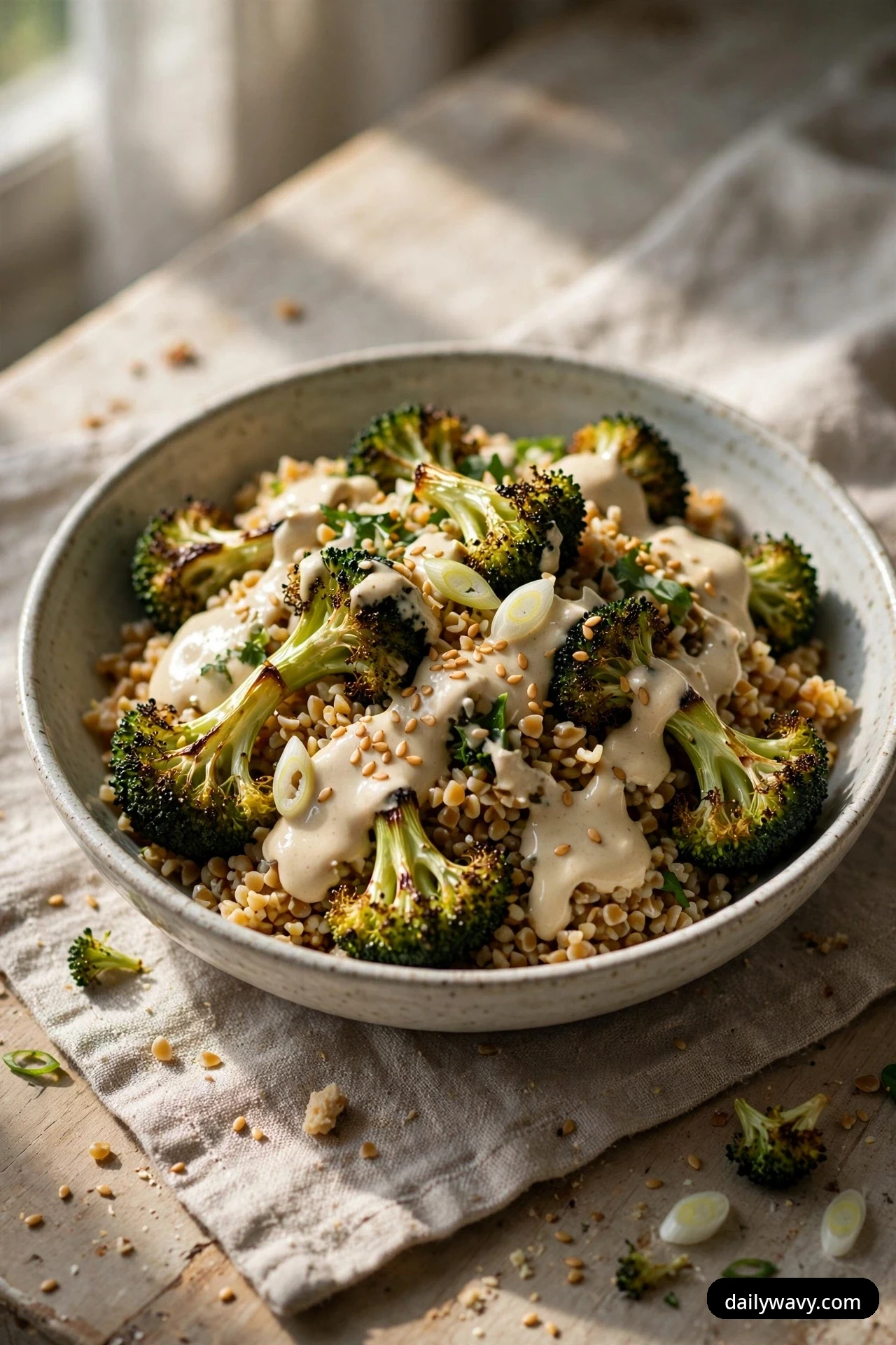 A closer view of a shallow bowl of bulgur salad topped with charred roasted broccoli florets, drizzled tahini dressing, toasted sesame seeds and sliced scallions on a linen cloth.
