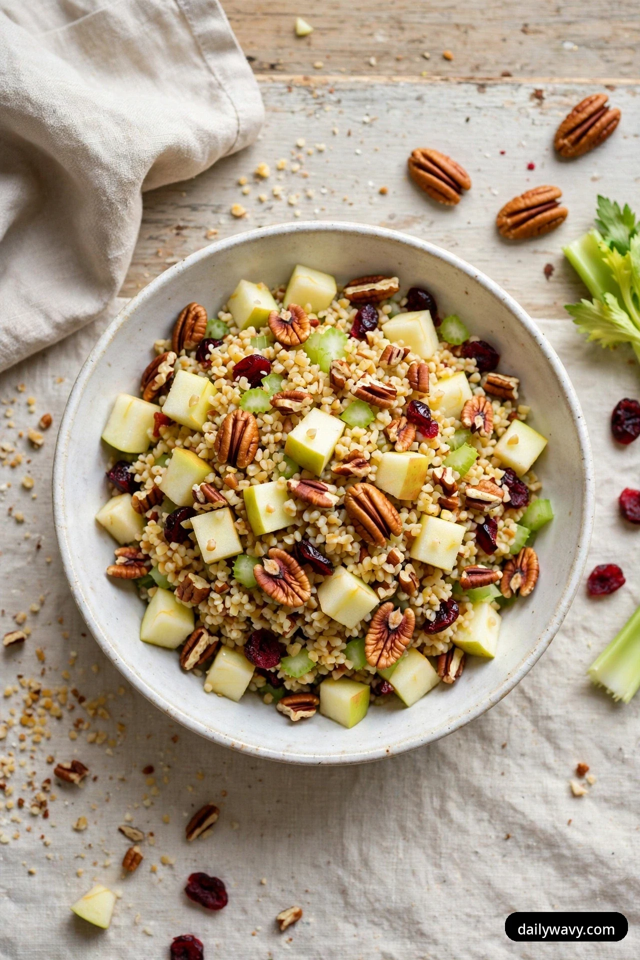 An overhead bowl of bulgur salad with diced apple, pecans, diced celery and dried cranberries sits on a linen cloth, with celery stalks beside it.