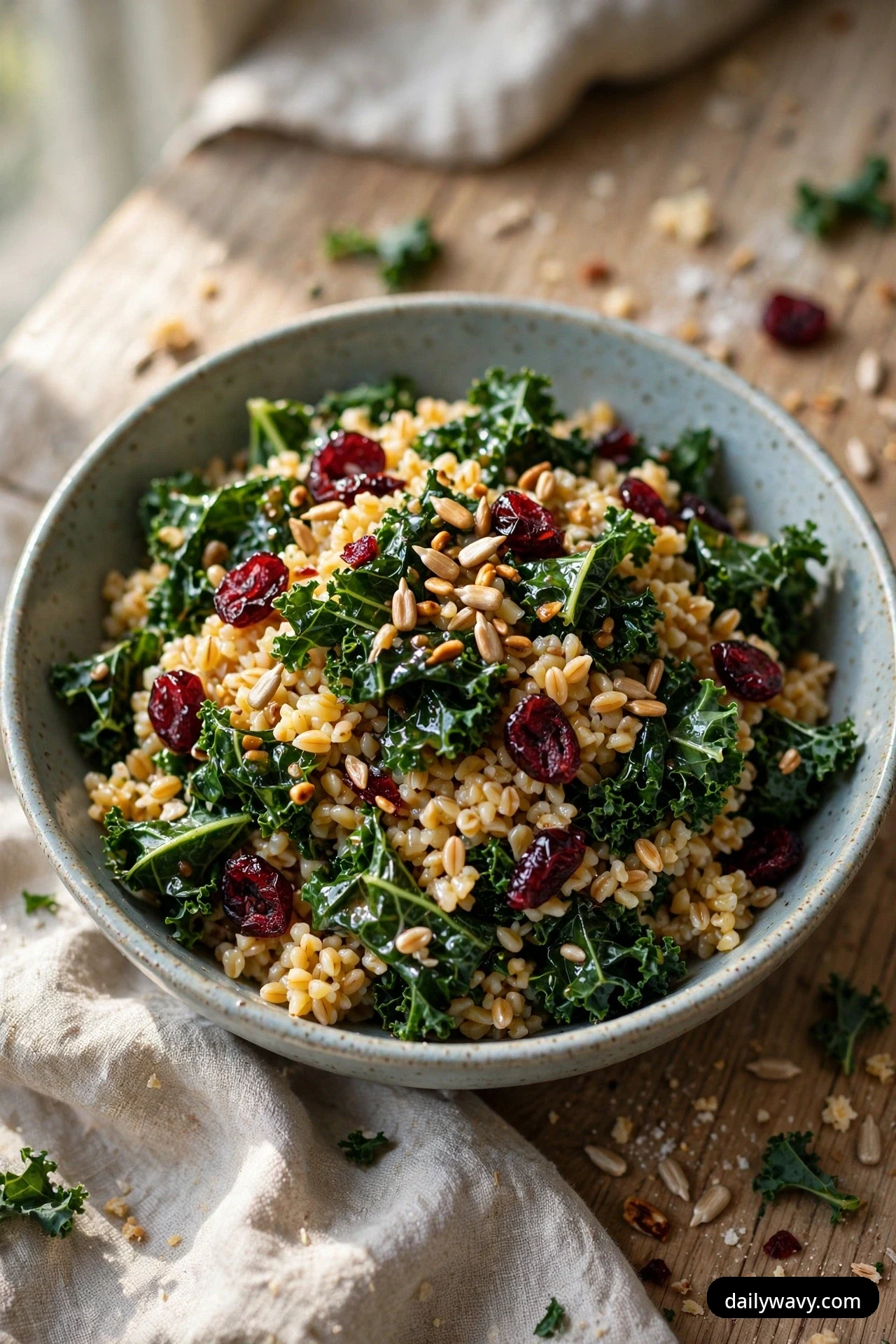 A bowl of bulgur salad with massaged kale leaves and dried cranberries is topped with toasted sunflower seeds on a wooden table.
