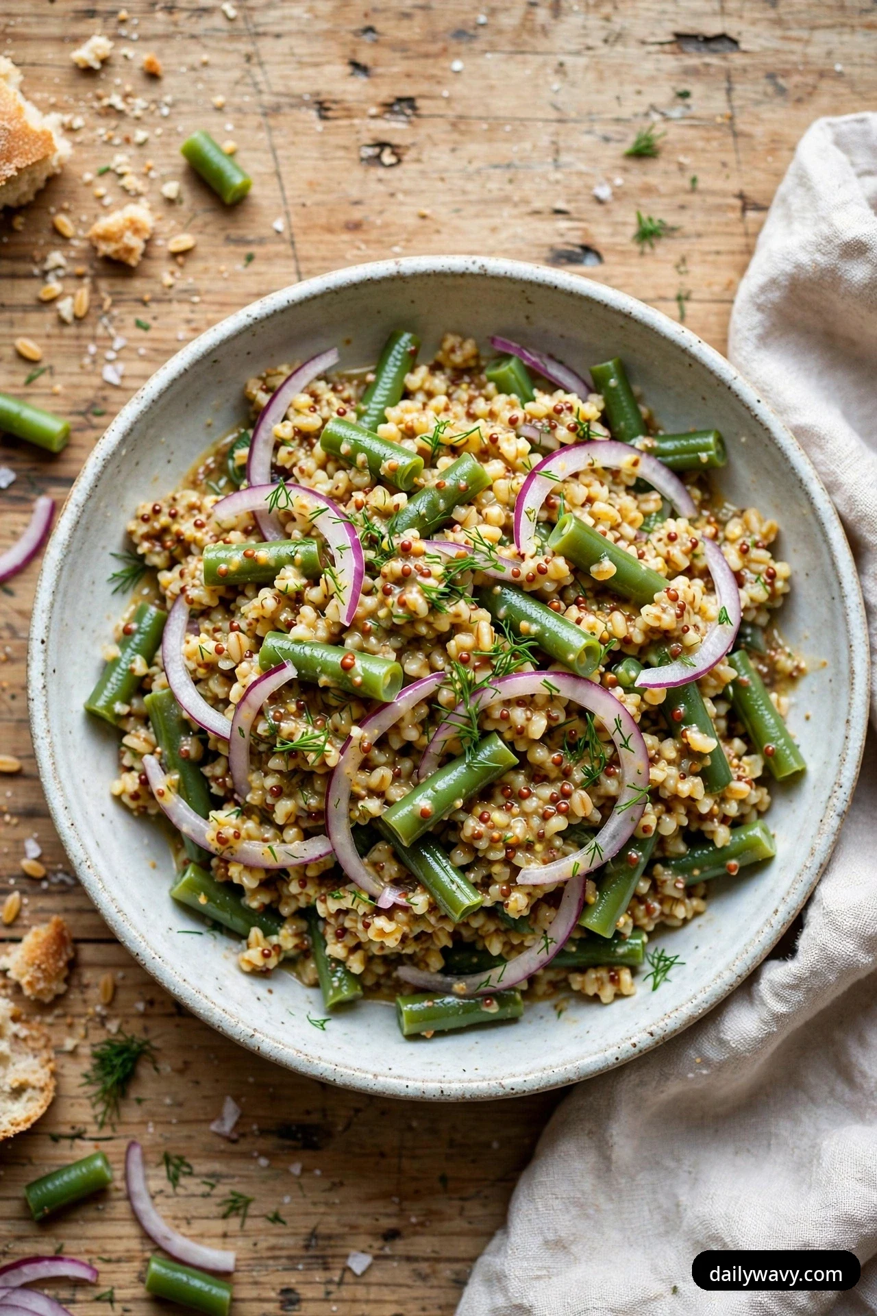 An overhead bowl of bulgur salad with blanched green bean pieces, red onion rings and fresh dill in a grainy mustard dressing rests on a rustic wooden table.