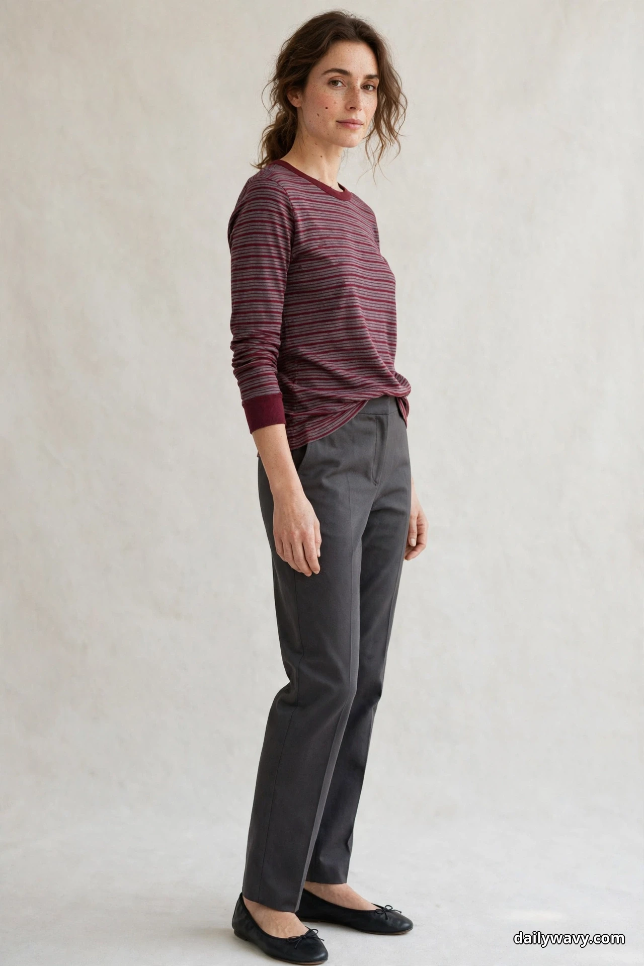 A woman stands in a three-quarter pose against a plain cream wall wearing a burgundy and gray thin-striped long-sleeve Breton top half-tucked into solid charcoal gray trousers, with black ballet flats