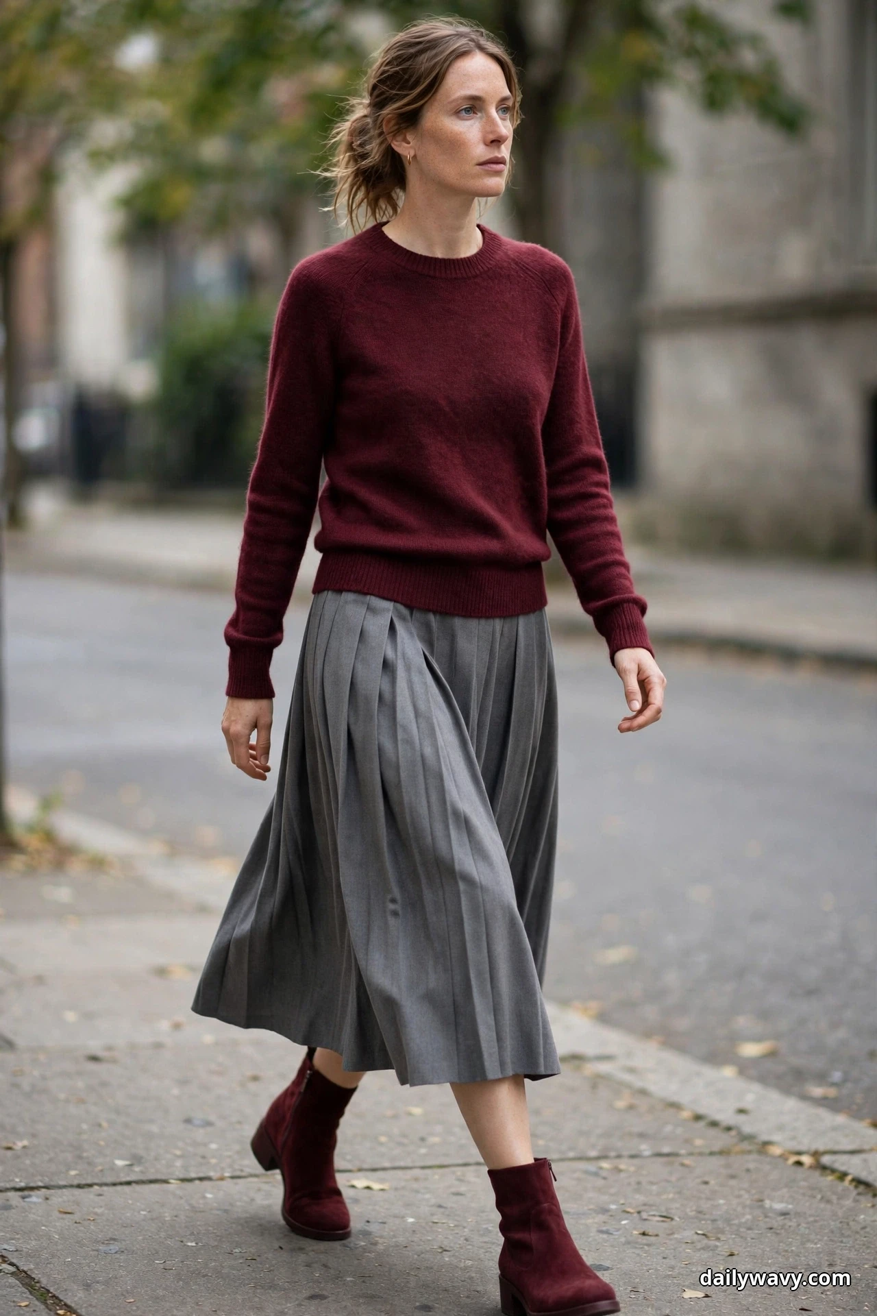 A woman walks along a city sidewalk wearing a burgundy fine-knit crewneck sweater over a gray pleated midi skirt, with burgundy suede ankle boots