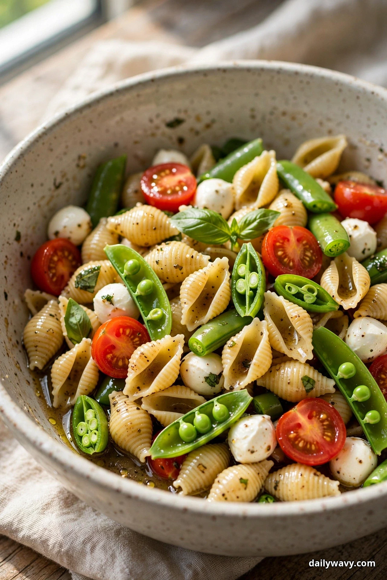 A close view of small shell pasta salad with halved sugar snap peas, mozzarella pearls, halved cherry tomatoes and basil fills a speckled bowl resting on linen