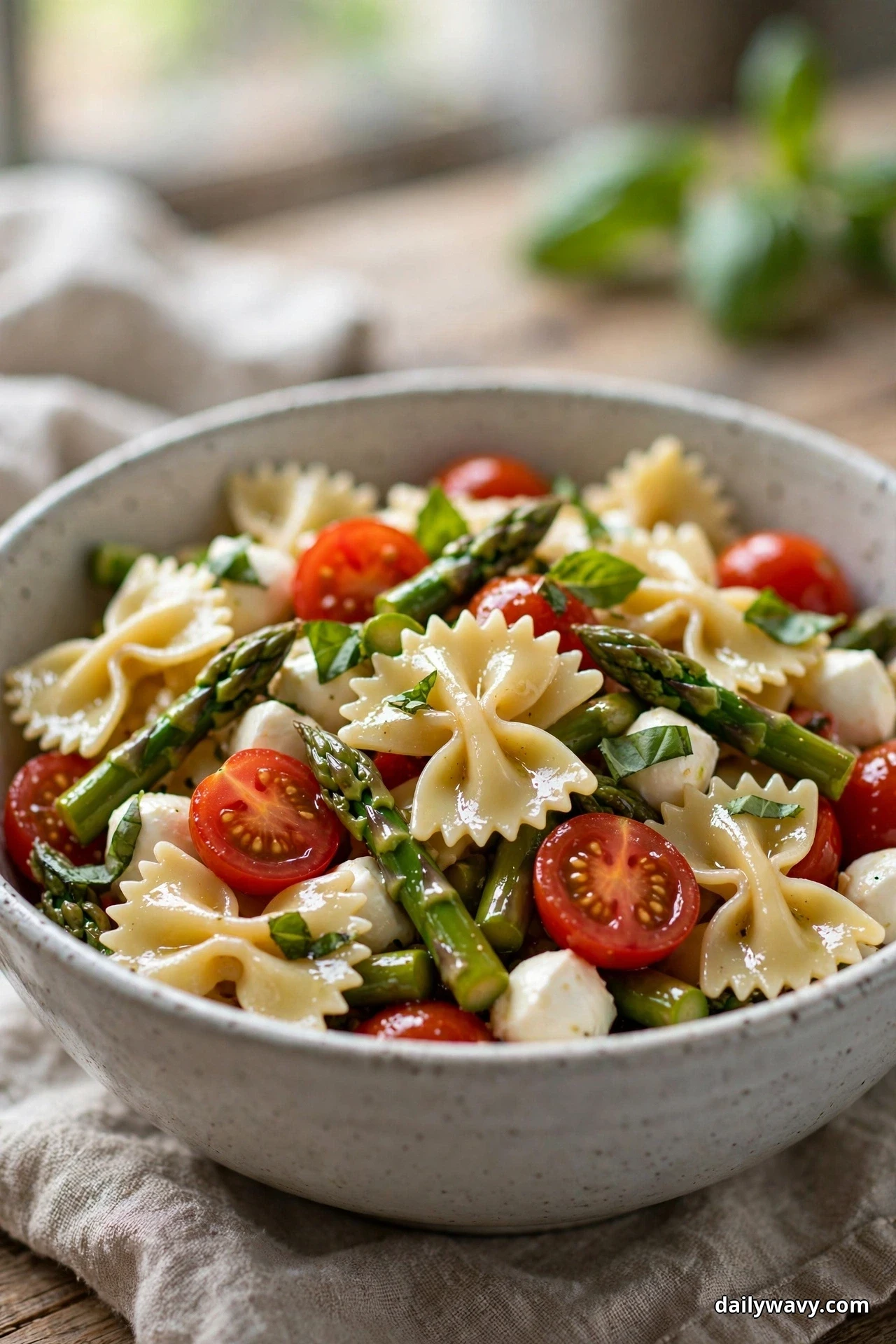 A bowl of farfalle pasta salad with asparagus pieces, mozzarella pearls, halved cherry tomatoes and torn basil sits on linen with blurred basil leaves behind it