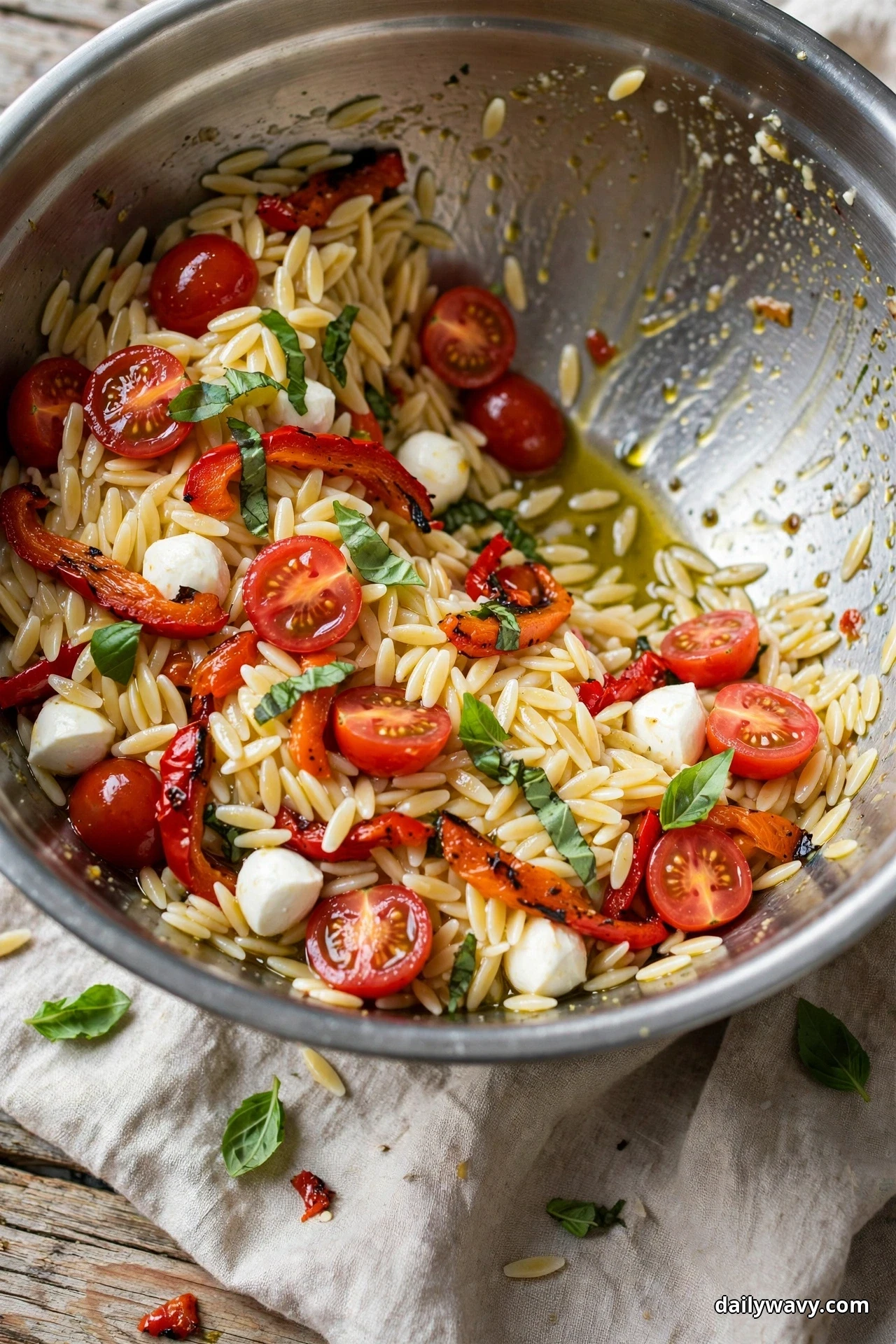A metal mixing bowl of orzo pasta salad with roasted red pepper strips, mozzarella pearls, halved cherry tomatoes and basil sits on linen with one portion scooped away