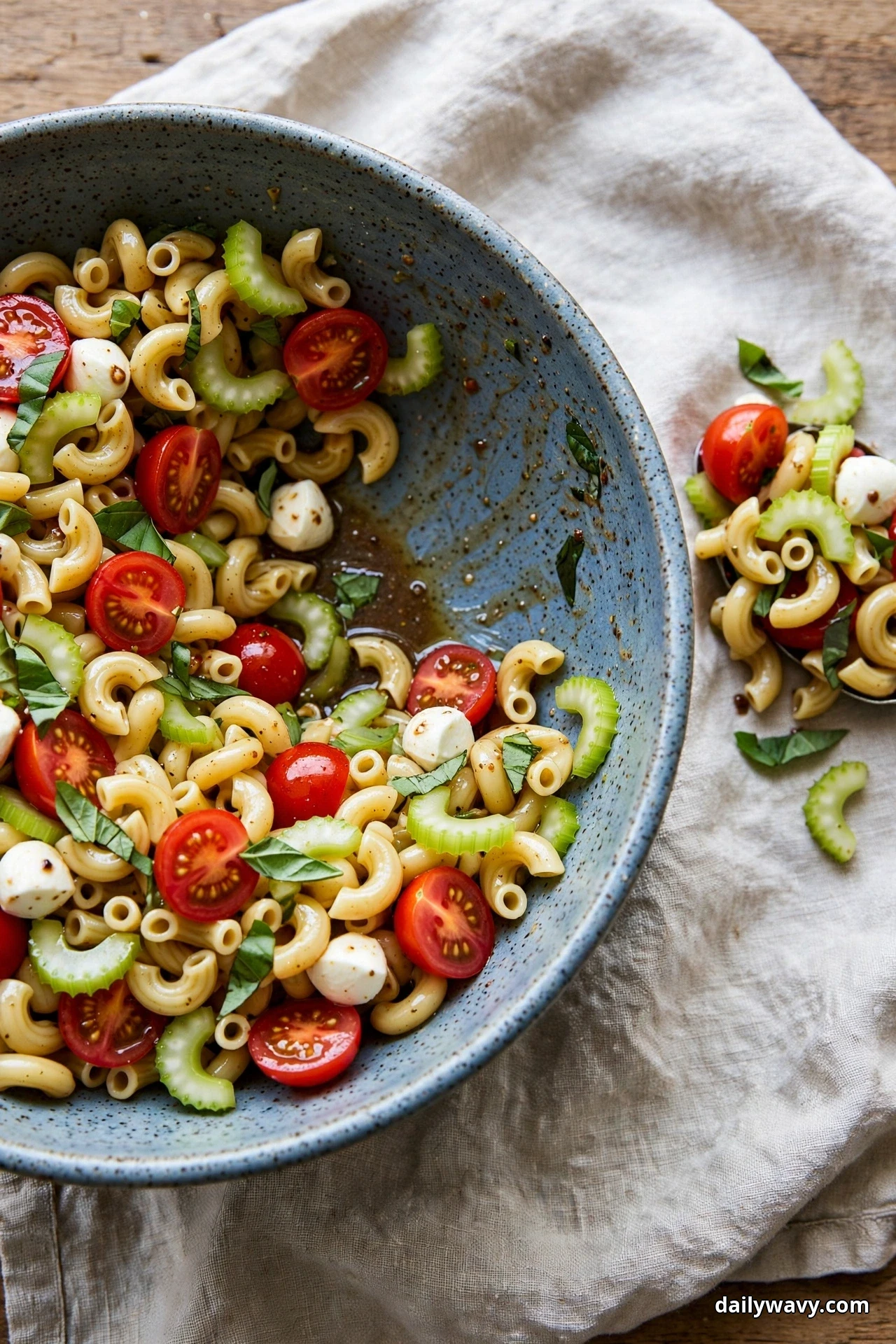 A blue-grey speckled bowl of elbow macaroni salad with sliced celery, mozzarella pearls, halved cherry tomatoes and basil rests on linen with a small serving spooned out beside it