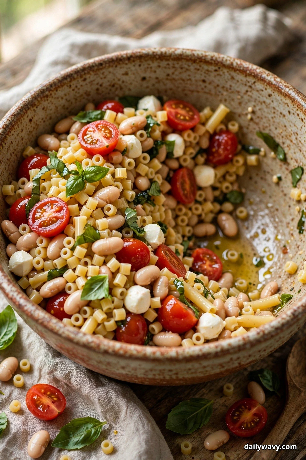 A rustic speckled bowl of ditalini pasta salad with cannellini beans, mozzarella pearls, halved cherry tomatoes and basil sits on a wooden table with a wooden spoon and scattered beans beside it