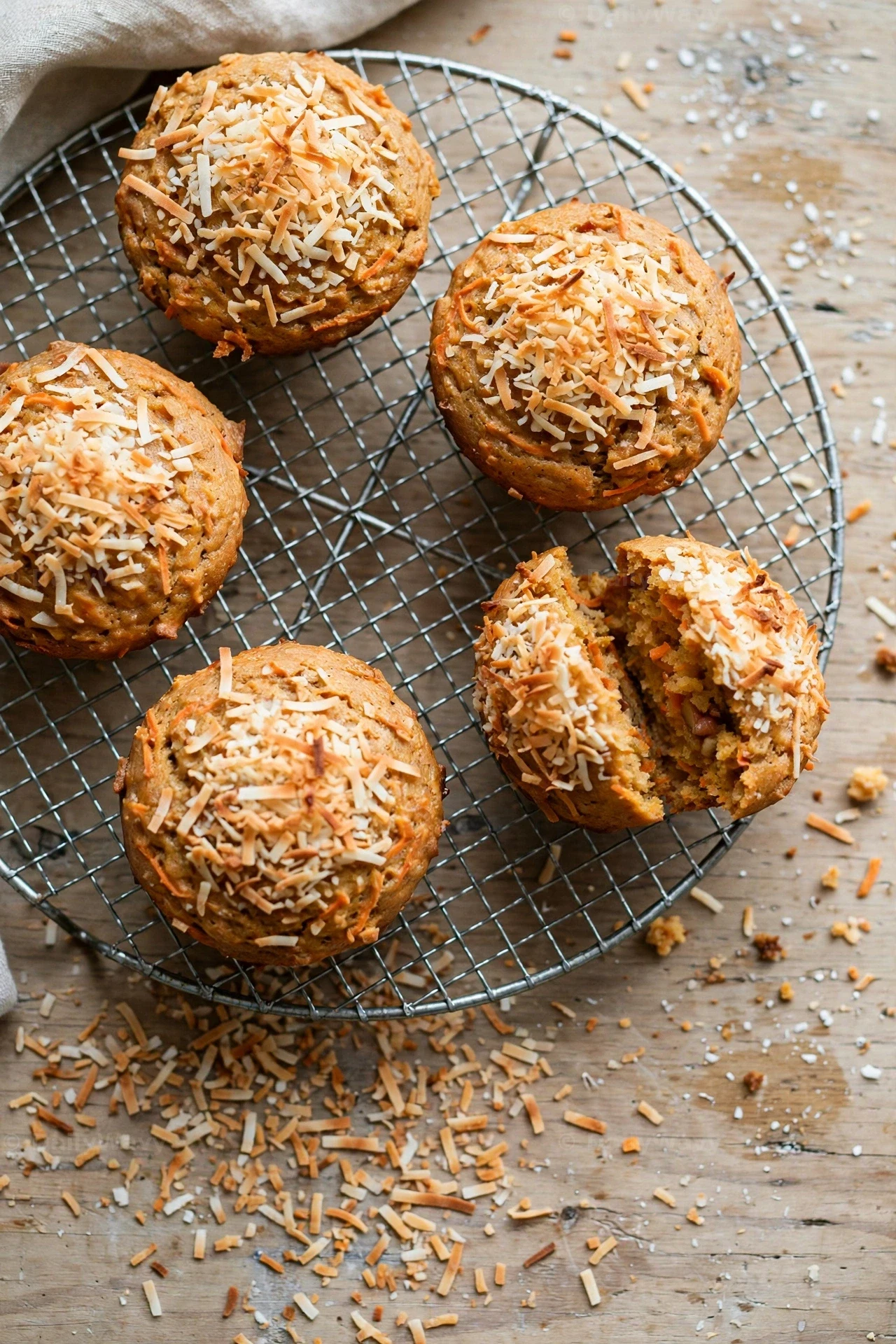 Toasted-coconut carrot muffins cool on a round wire rack seen from above, and one muffin is split open among scattered coconut flakes.