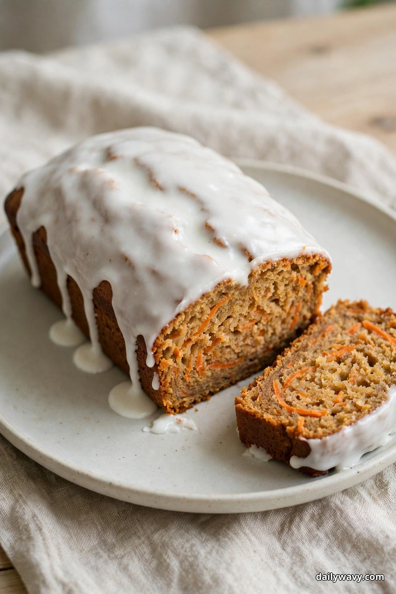 A carrot quick bread loaf covered in white yogurt glaze dripping down its sides sits on a ceramic plate, with one slice beside it showing orange carrot shreds in the crumb