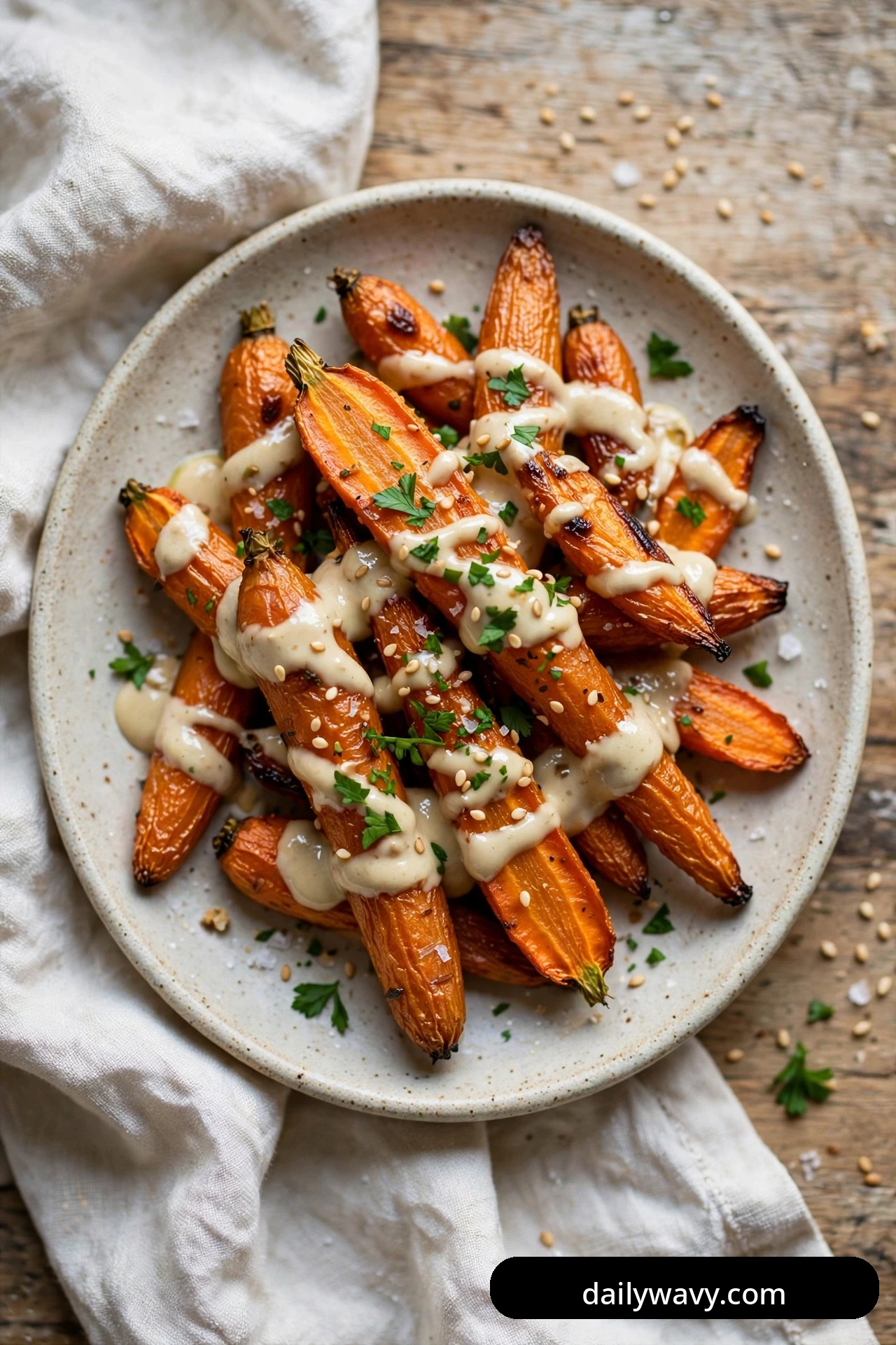Long roasted carrots on a speckled plate are drizzled with creamy tahini sauce and sprinkled with sesame seeds and chopped parsley.