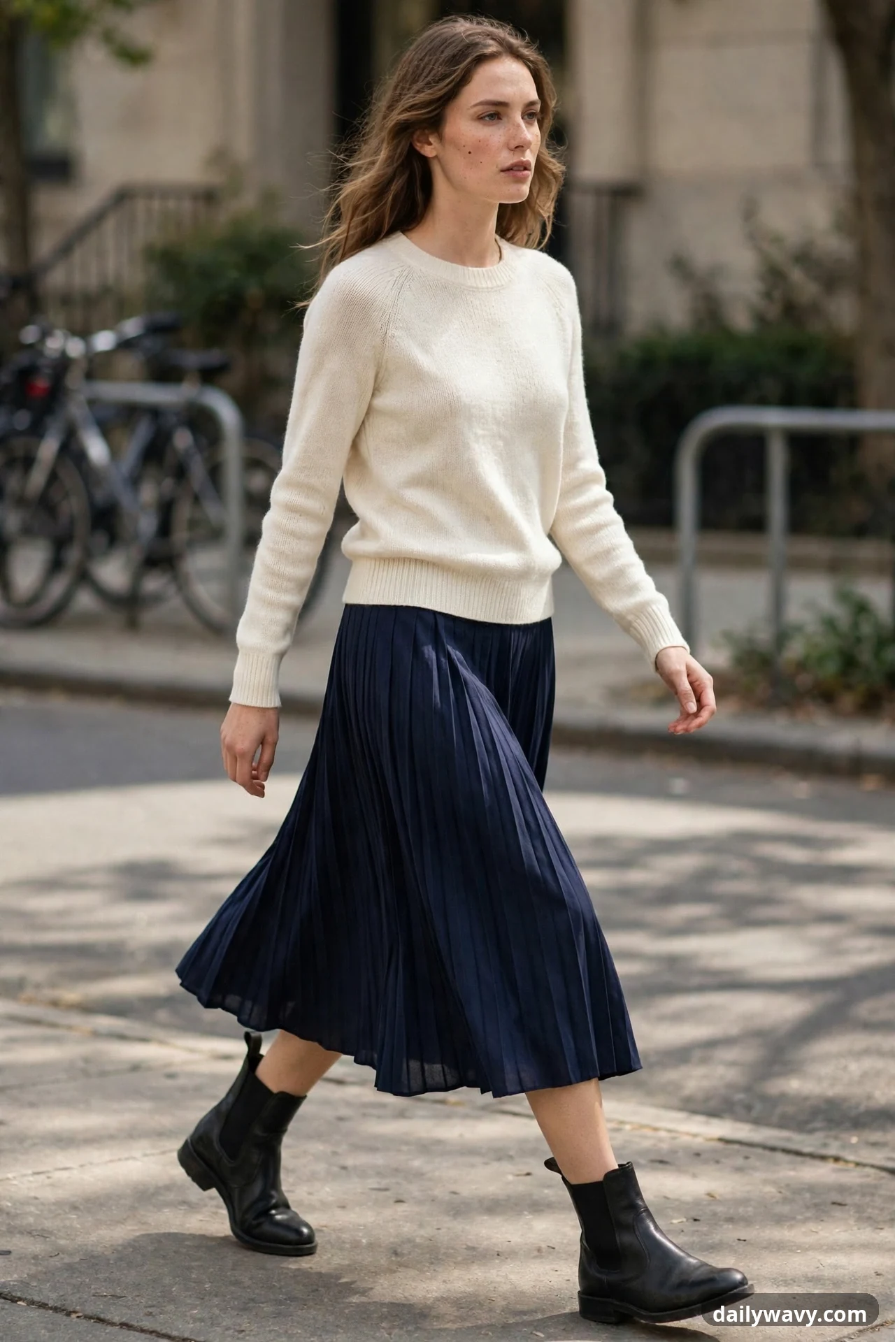 A woman walks in a cream crewneck sweater and navy pleated midi skirt, with bare legs above black leather chelsea boots with elastic side panels.