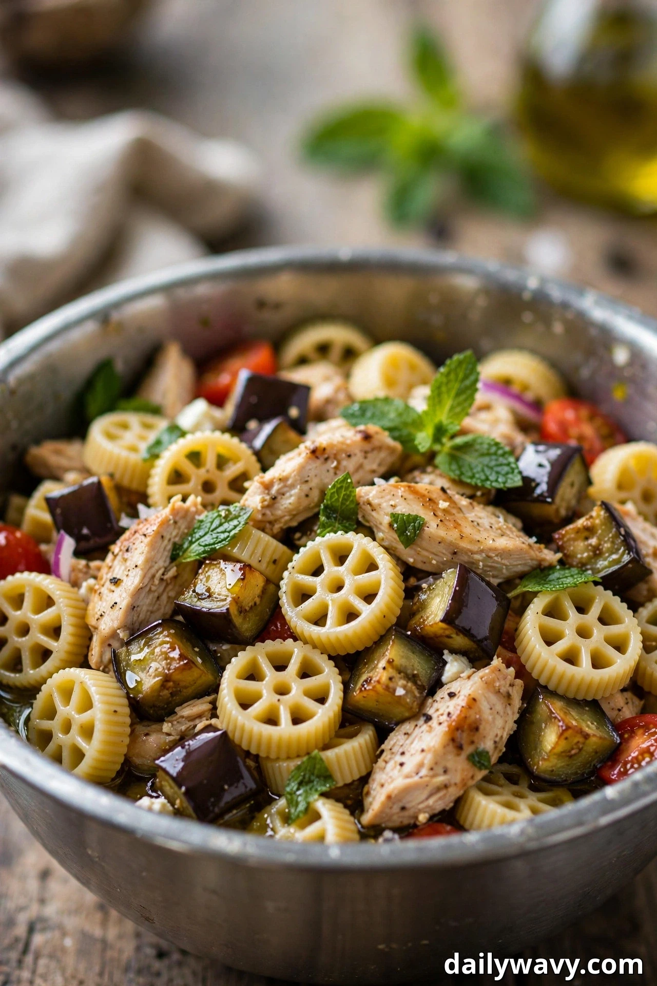 A metal bowl holds rotelle pasta salad tossed with roasted eggplant cubes, sliced chicken and fresh mint leaves in a glossy olive oil dressing.