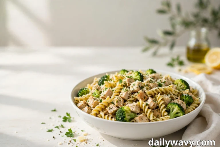 A white bowl of twisted pasta salad holds diced chicken, broccoli florets, grated Parmesan and chopped parsley, with a small bottle of olive oil and lemon halves blurred in the bright background
