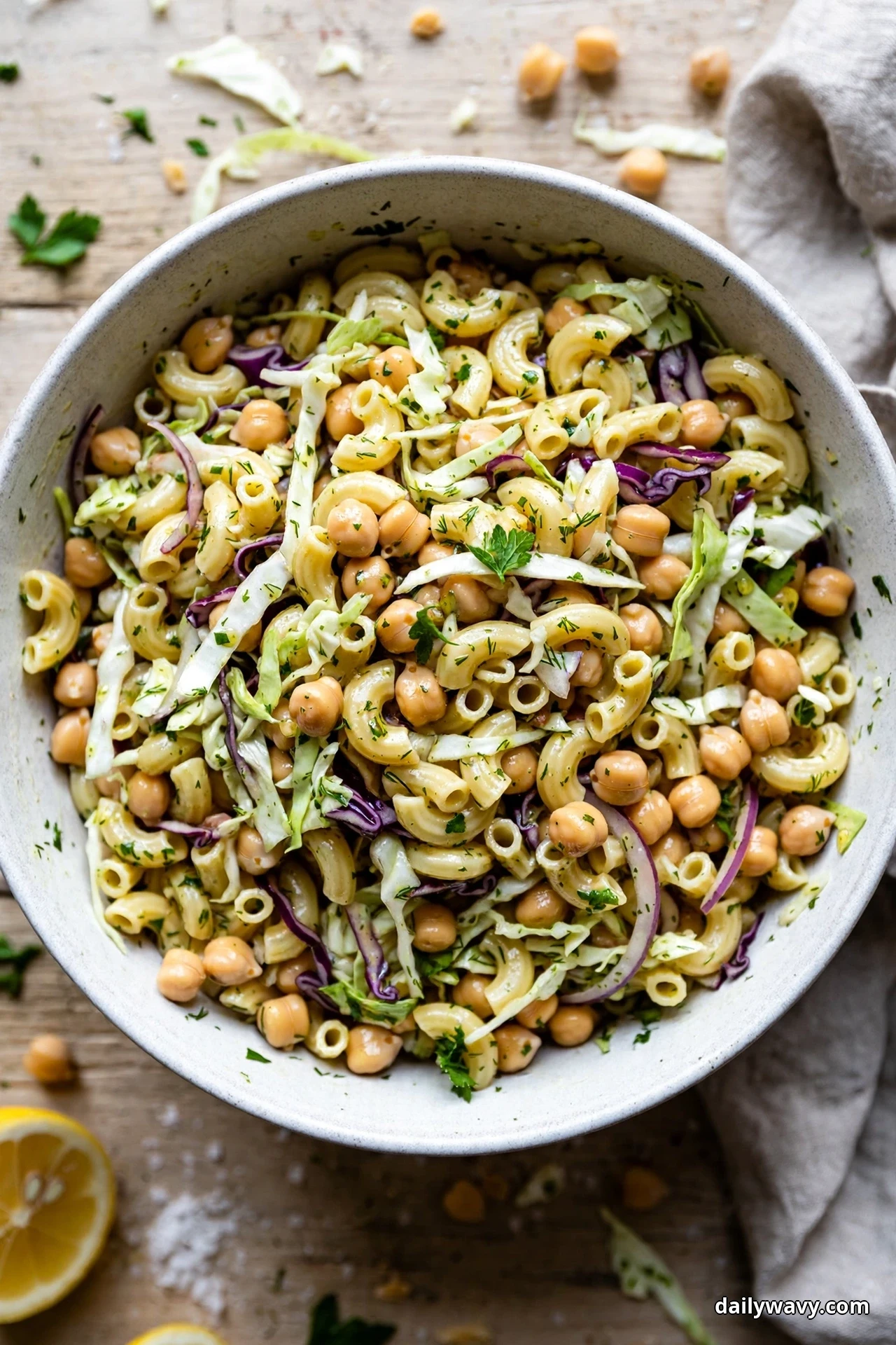 An overhead view of a speckled bowl of elbow pasta tossed with chickpeas and thinly shredded cabbage in a mustard vinaigrette rests on a wooden table