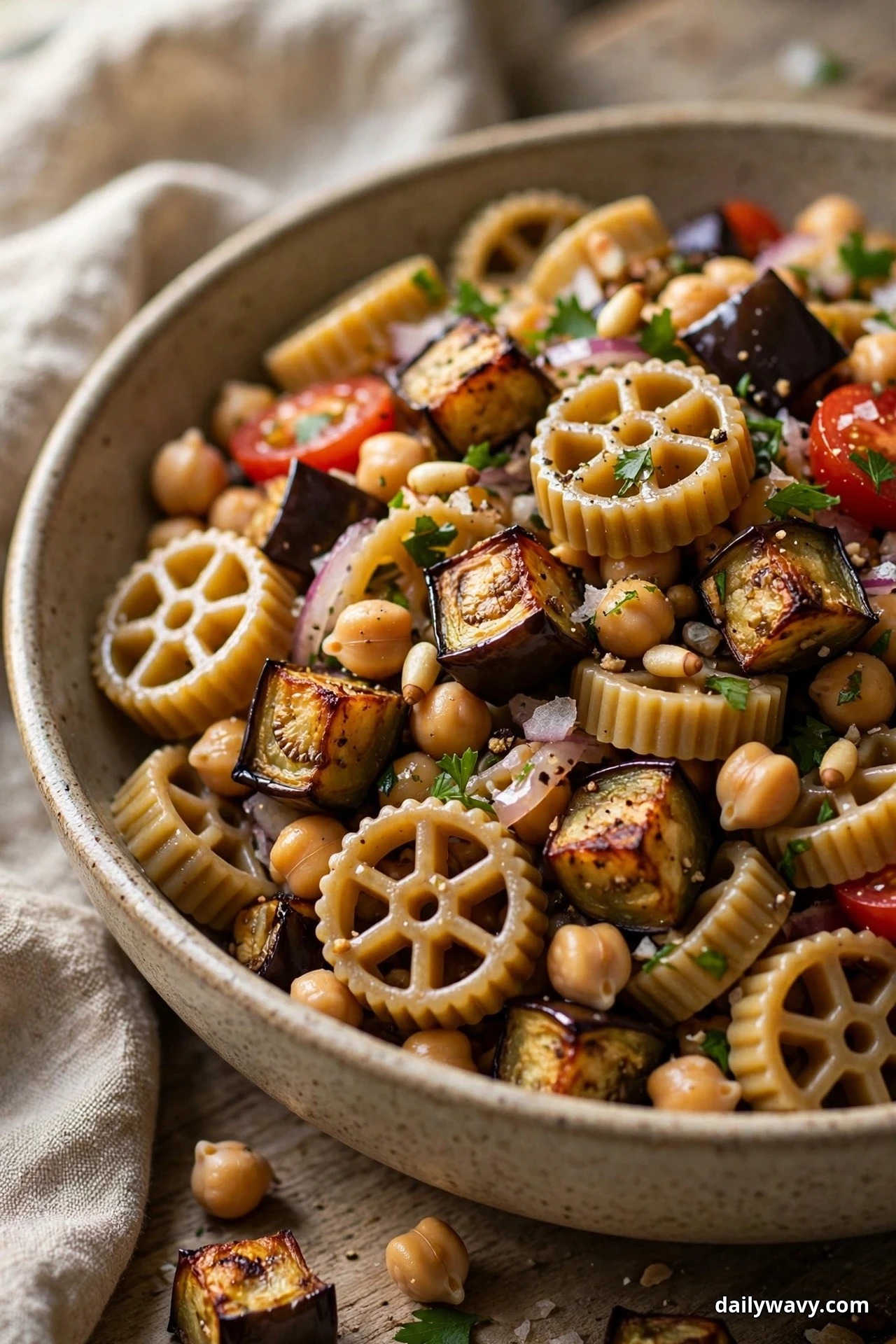 A close view of rotelle wagon wheels tossed with golden roasted eggplant cubes, chickpeas and halved cherry tomatoes fills a speckled bowl over linen