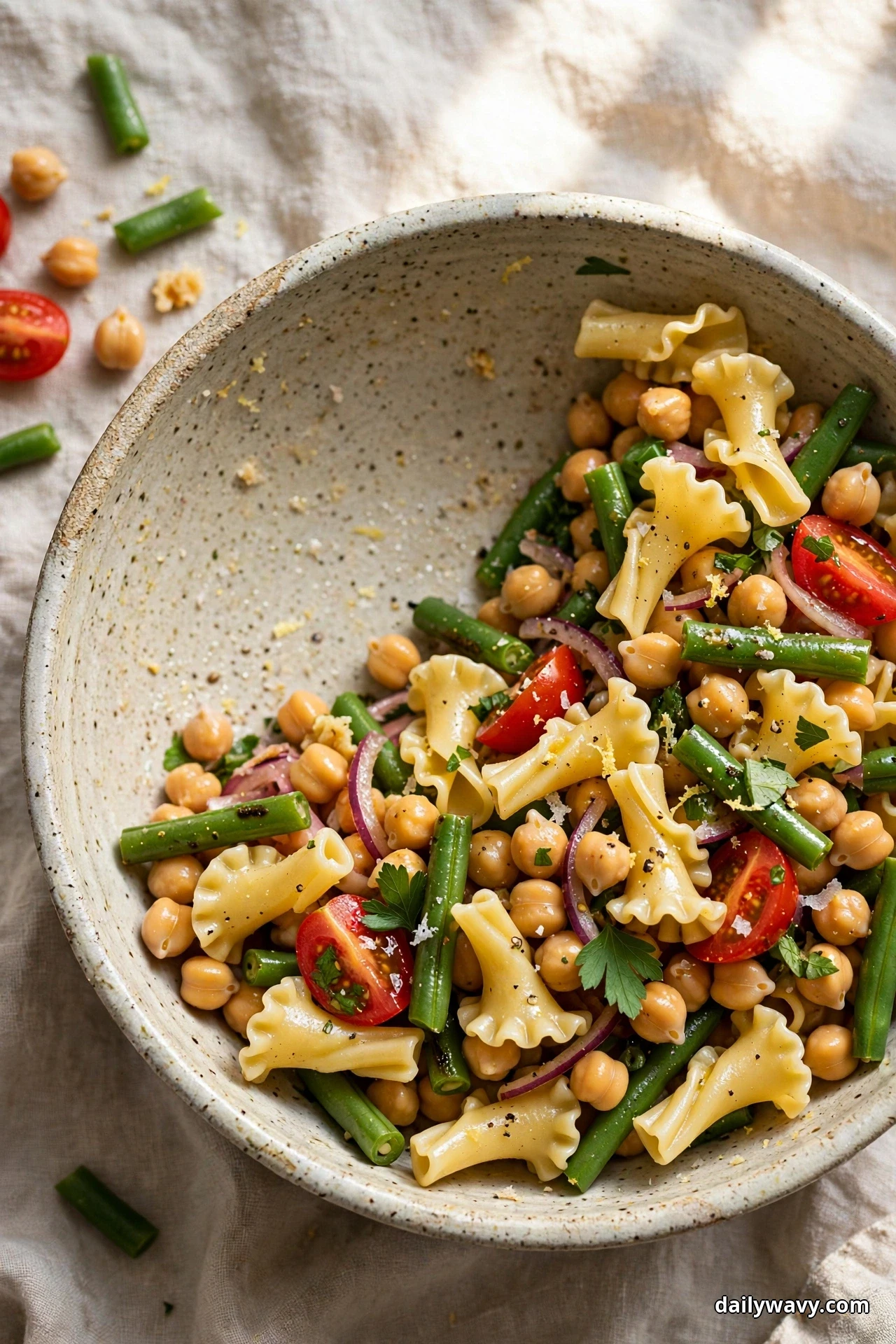 A speckled bowl of campanelle pasta tossed with chickpeas and green bean pieces in a lemon dressing sits on linen with one serving removed to show the bowl interior