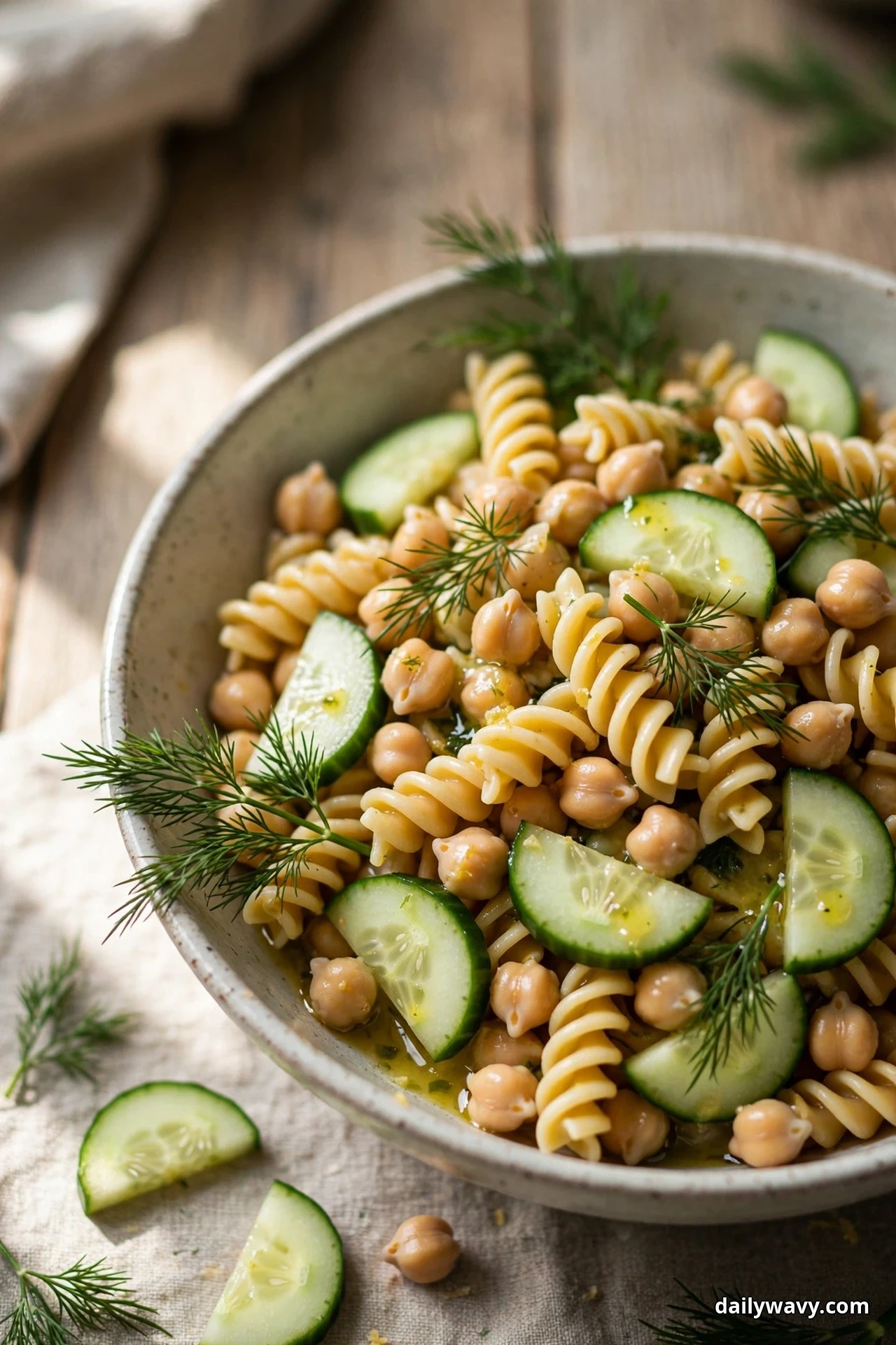 A bowl of rotini spirals tossed with chickpeas and cucumber half moons is topped with fresh dill fronds and sits over linen on a wooden table
