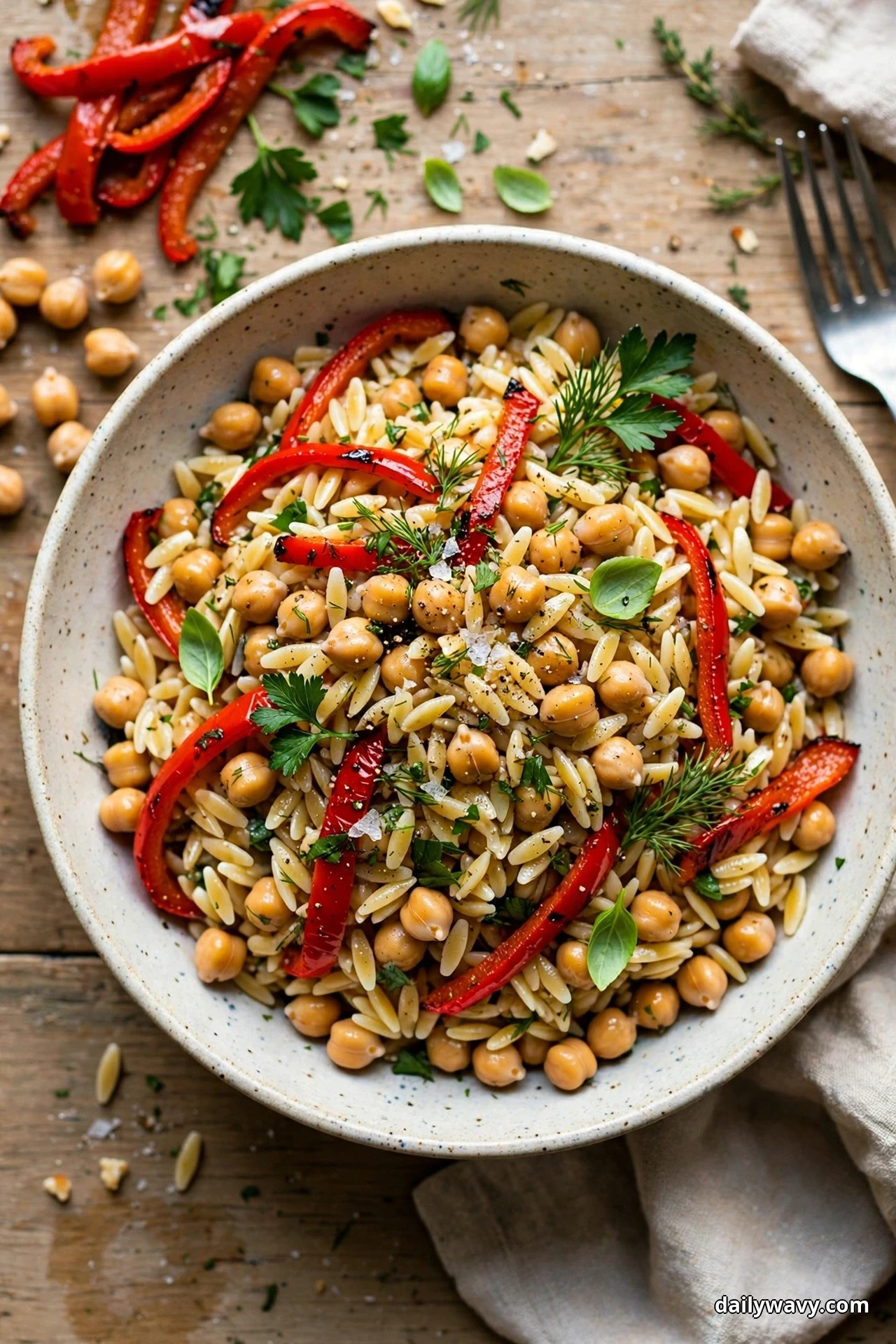 An overhead view of a speckled bowl of orzo tossed with chickpeas and roasted red pepper strips sits on a wooden table with extra pepper strips and chickpeas scattered beside it