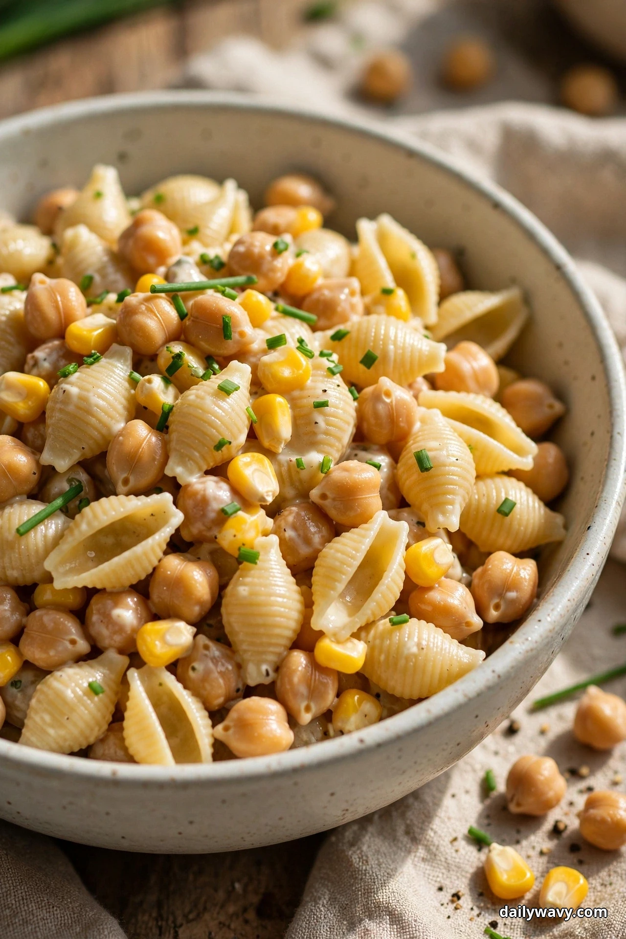 A close view of medium pasta shells tossed with chickpeas and corn kernels in a creamy dressing is sprinkled with snipped chives in a speckled bowl