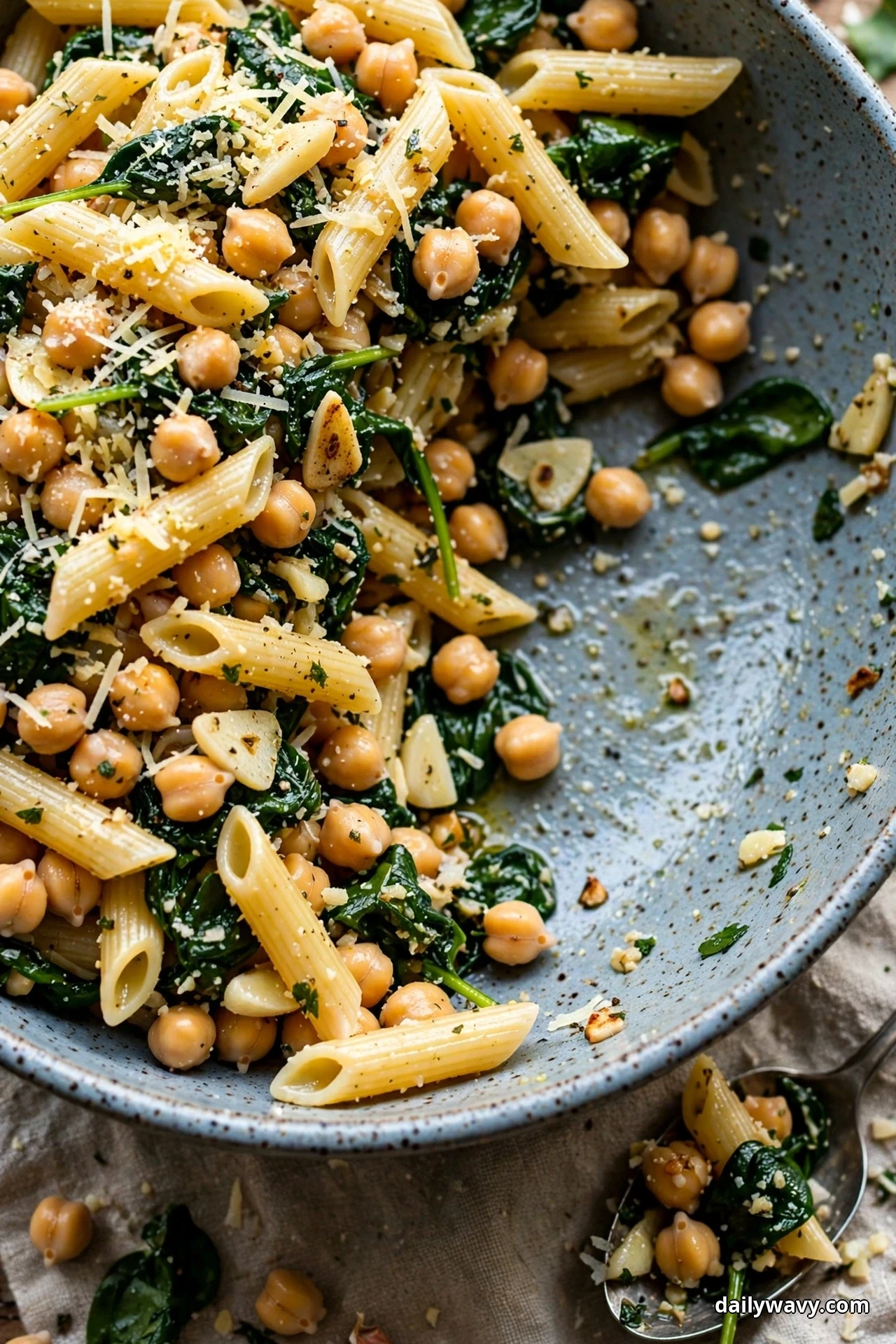 A blue speckled bowl of penne tossed with chickpeas, wilted spinach and sliced garlic is topped with grated parmesan, with one serving spooned out beside the bowl
