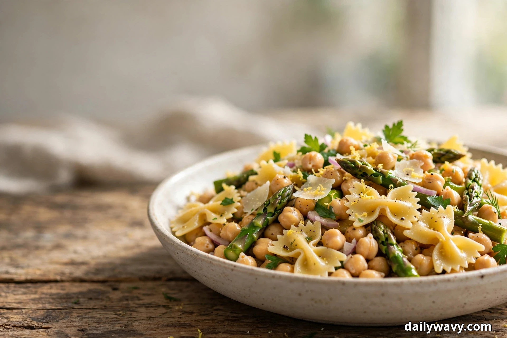 A shallow speckled bowl of farfalle bow ties tossed with chickpeas, asparagus spears, shaved parmesan and lemon zest sits on a rustic wooden table with linen behind it