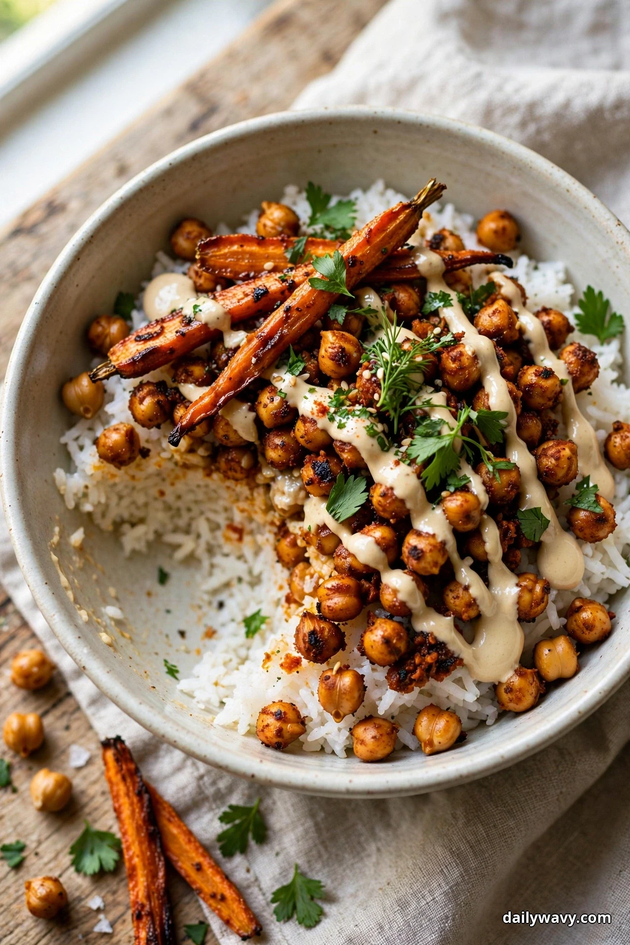 A bowl of white rice topped with harissa-roasted carrots, spiced chickpeas and zigzags of tahini sauce is garnished with parsley leaves, with a serving already scooped out