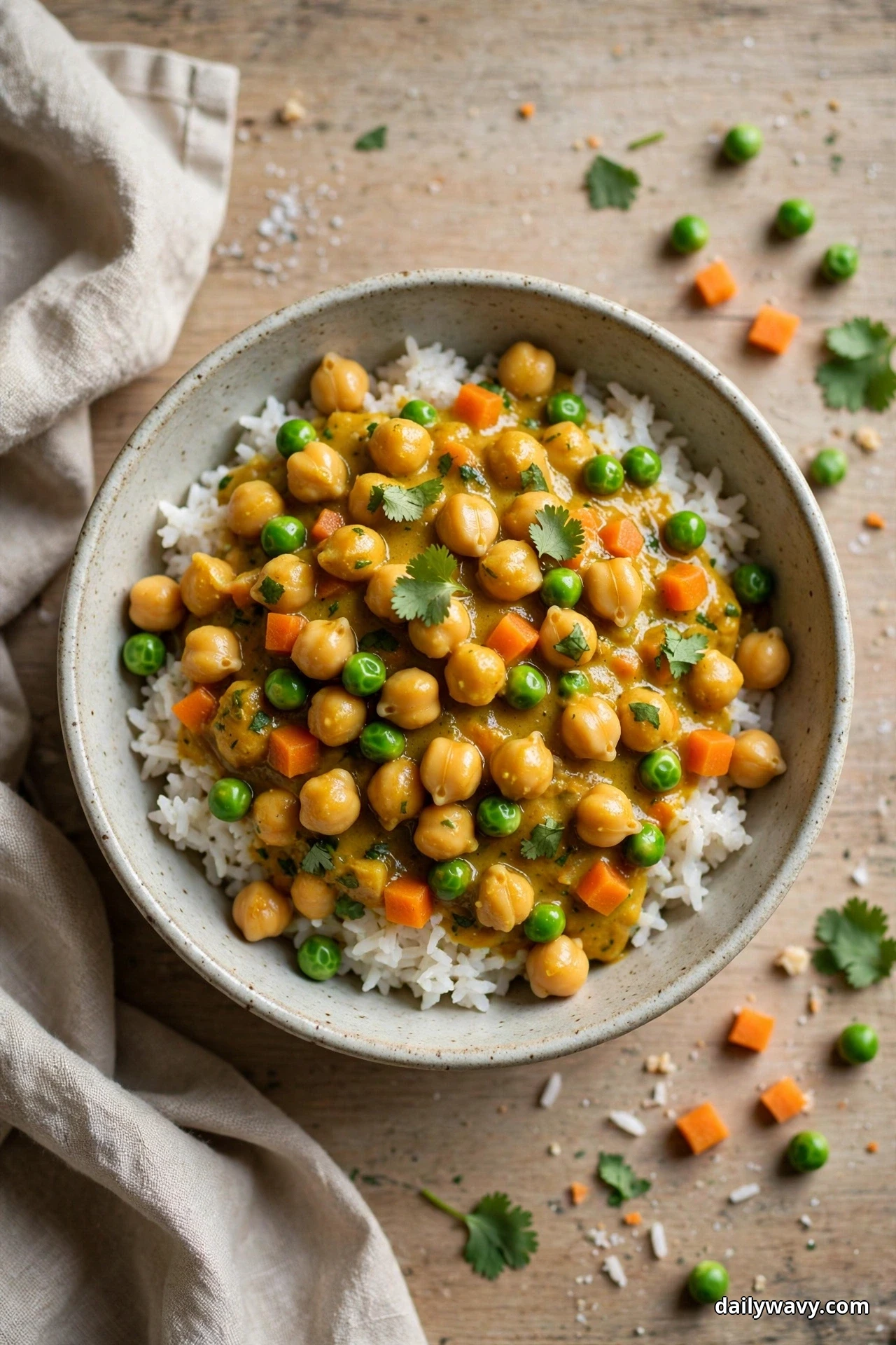 An overhead bowl of white rice topped with chickpeas, peas and diced carrot in a golden curry sauce is garnished with cilantro leaves