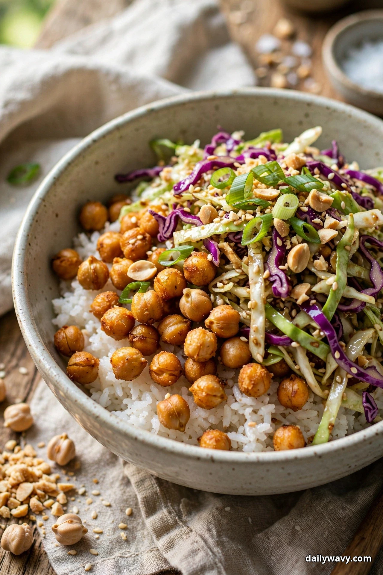 A bowl of white rice topped with roasted chickpeas and a heap of green and purple cabbage slaw is finished with sliced scallions, crushed peanuts and sesame seeds