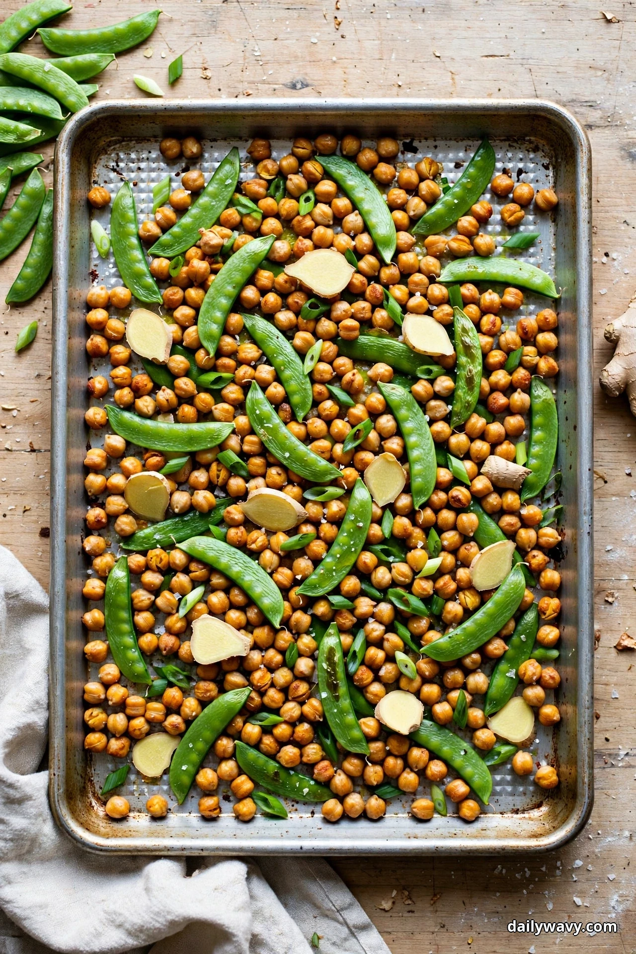 Seen from above, a sheet pan of roasted chickpeas, whole sugar snap peas, sliced fresh ginger and chopped green onions rests on a wooden table
