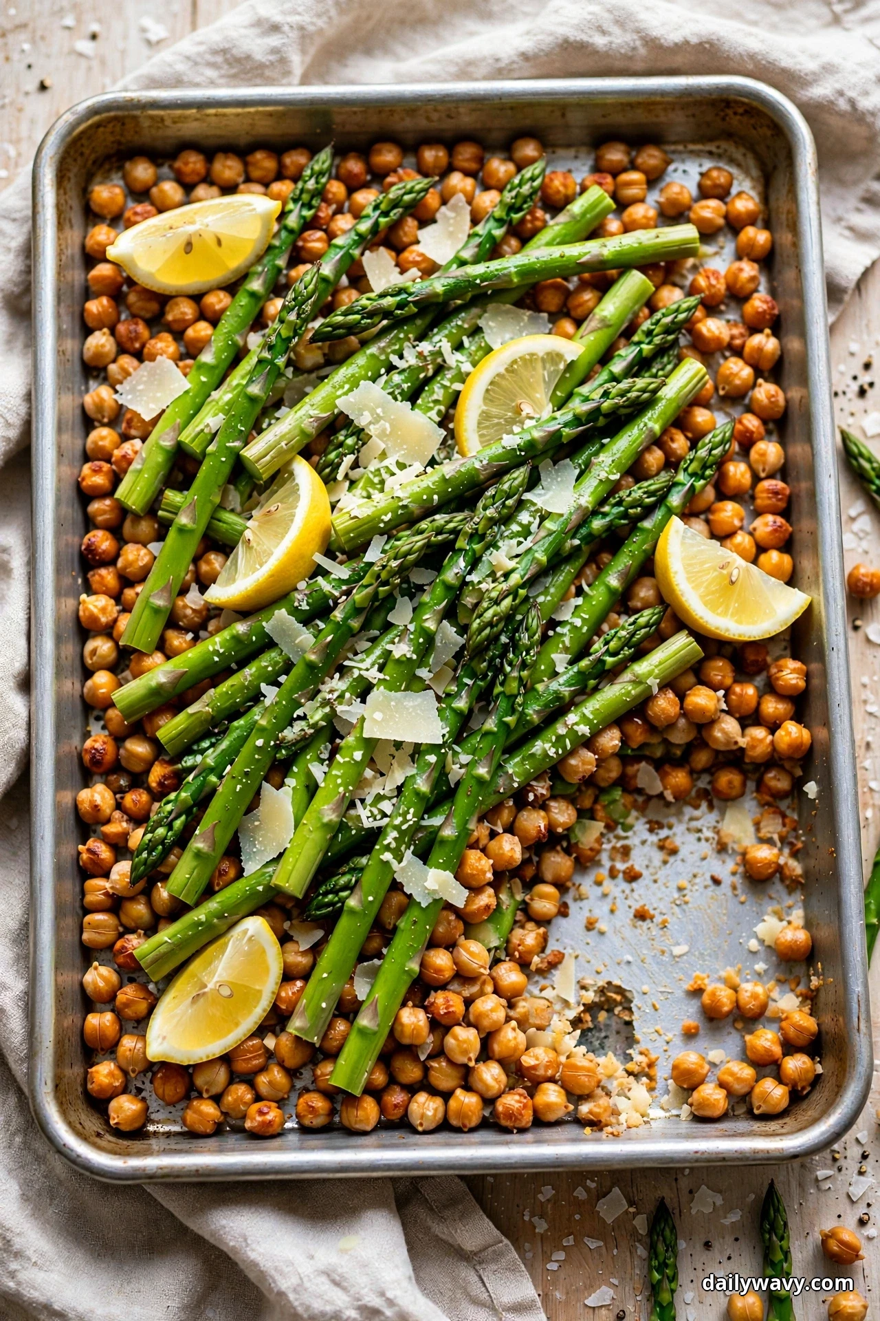 Green asparagus spears lie across roasted chickpeas with lemon wedges and Parmesan shavings on a sheet pan with one serving removed