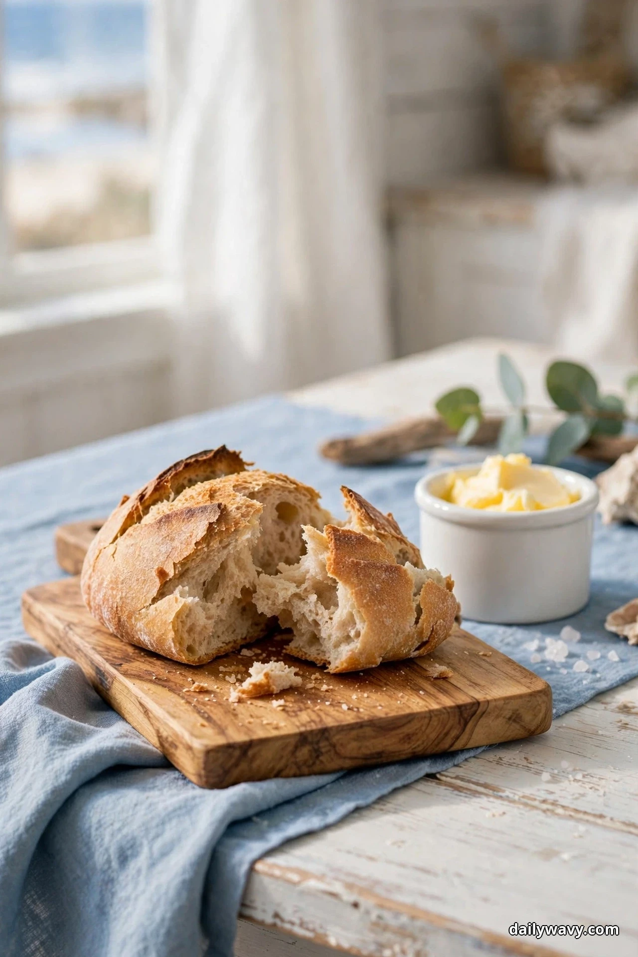 A torn rustic loaf rests on a small wooden bread board beside a white crock of butter on a coastal table dressed with soft blue linen