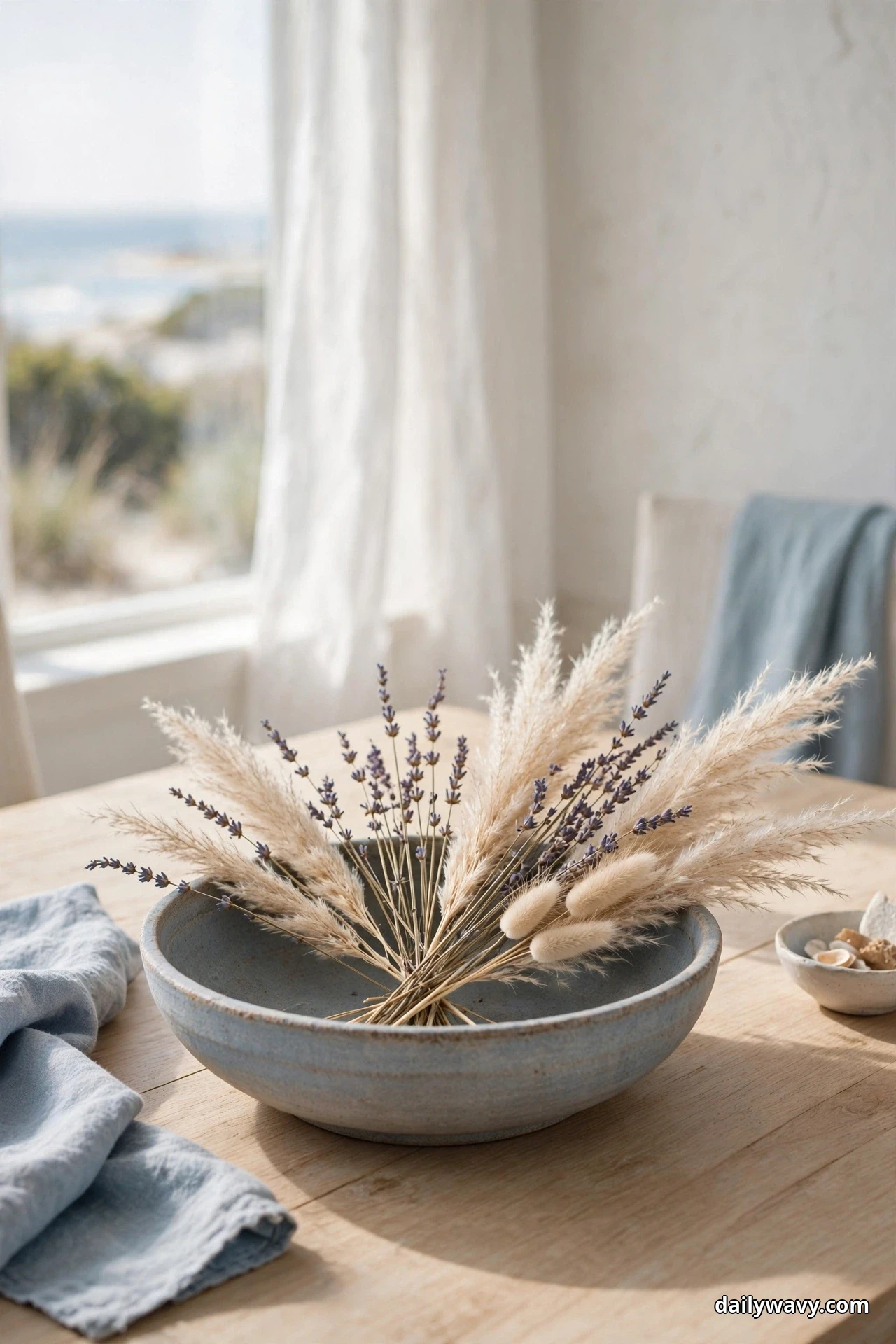 A low blue-gray stoneware bowl holds a loose fan of dried pampas grass, lavender and bunny tails on a light wood table