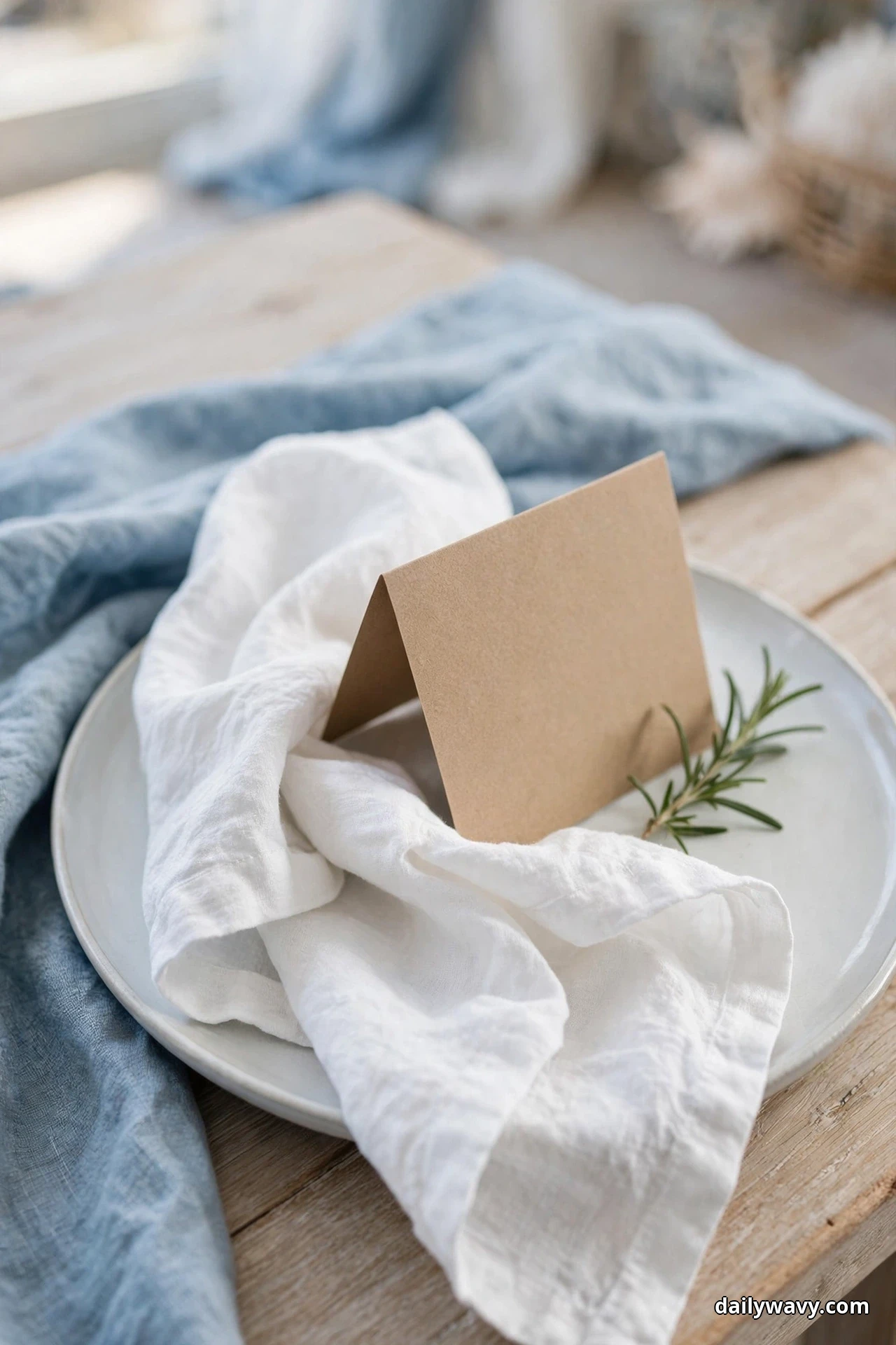 A white plate holds a folded white linen napkin with a blank kraft tent card leaning against it and a sprig of rosemary beside it