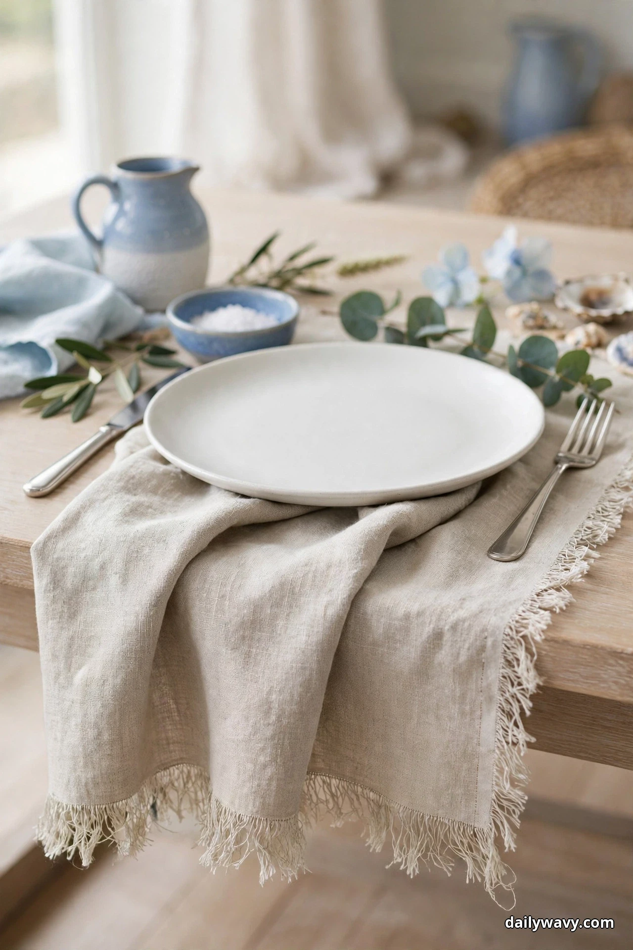 A white dinner plate sits on a fringed oatmeal linen strip running in from the table edge, with a fork at its left and eucalyptus sprigs behind it