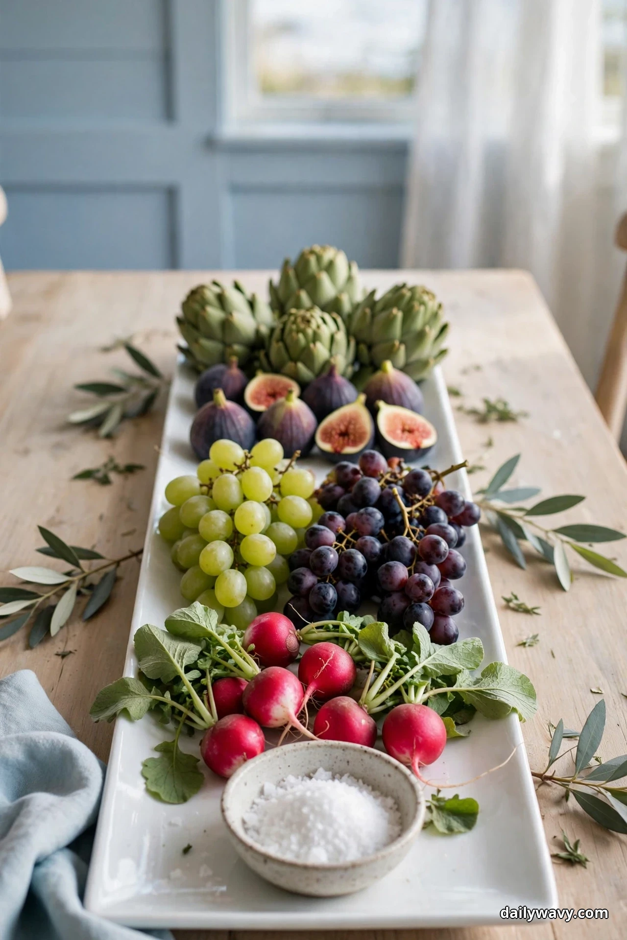 A long white platter is arranged with artichokes, figs, green and purple grapes and radishes, with a small bowl of flaky salt at one end
