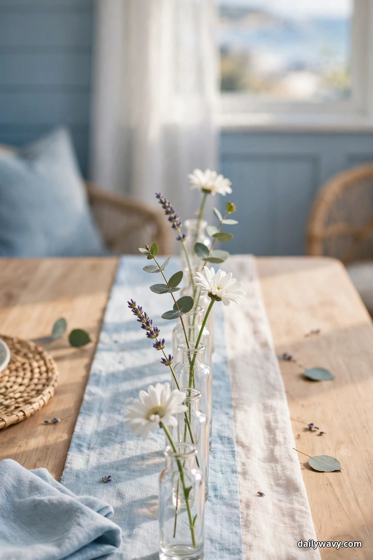 Small clear bud vases holding single stems of white daisies, lavender and eucalyptus stand along a soft blue striped runner on a wood table