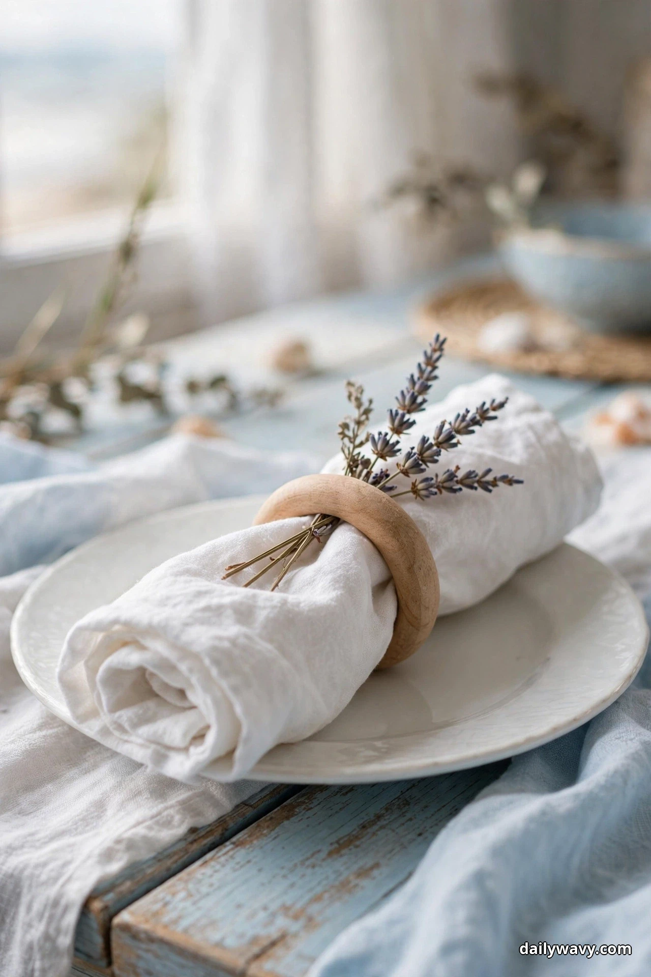 A rolled white linen napkin held by a plain wooden ring with sprigs of dried lavender through it rests on a white plate