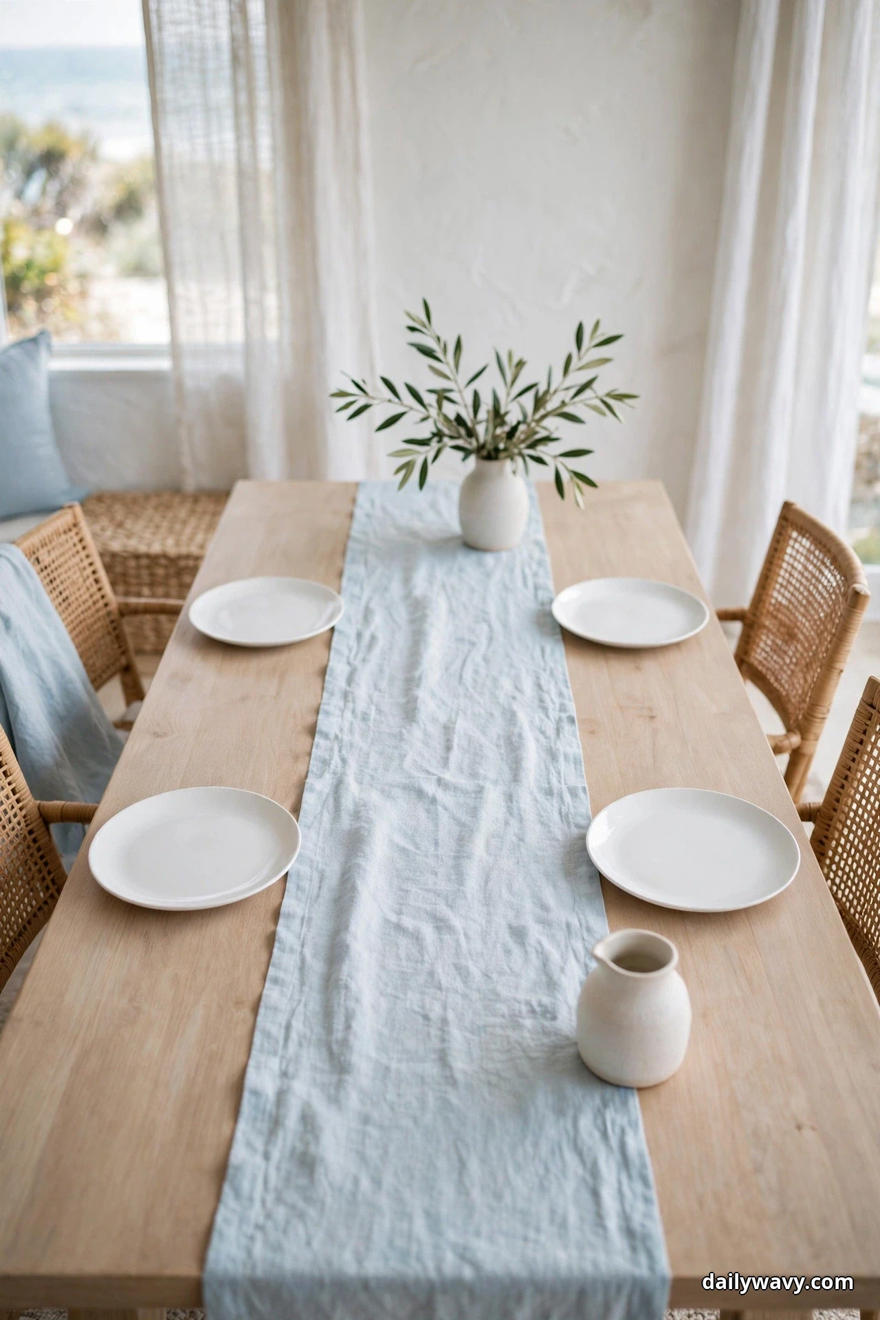 A narrow pale blue linen runner runs down the center of a light wood table set with plain white plates and a small white vase of olive sprigs