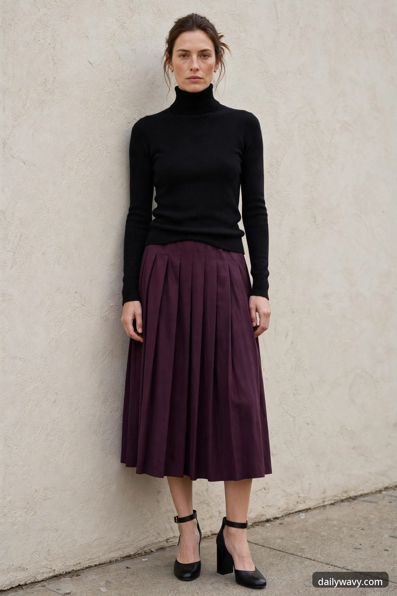 A woman stands against a plain stucco wall wearing a black turtleneck knit, a plum pleated midi skirt and black ankle-strap block-heel pumps.
